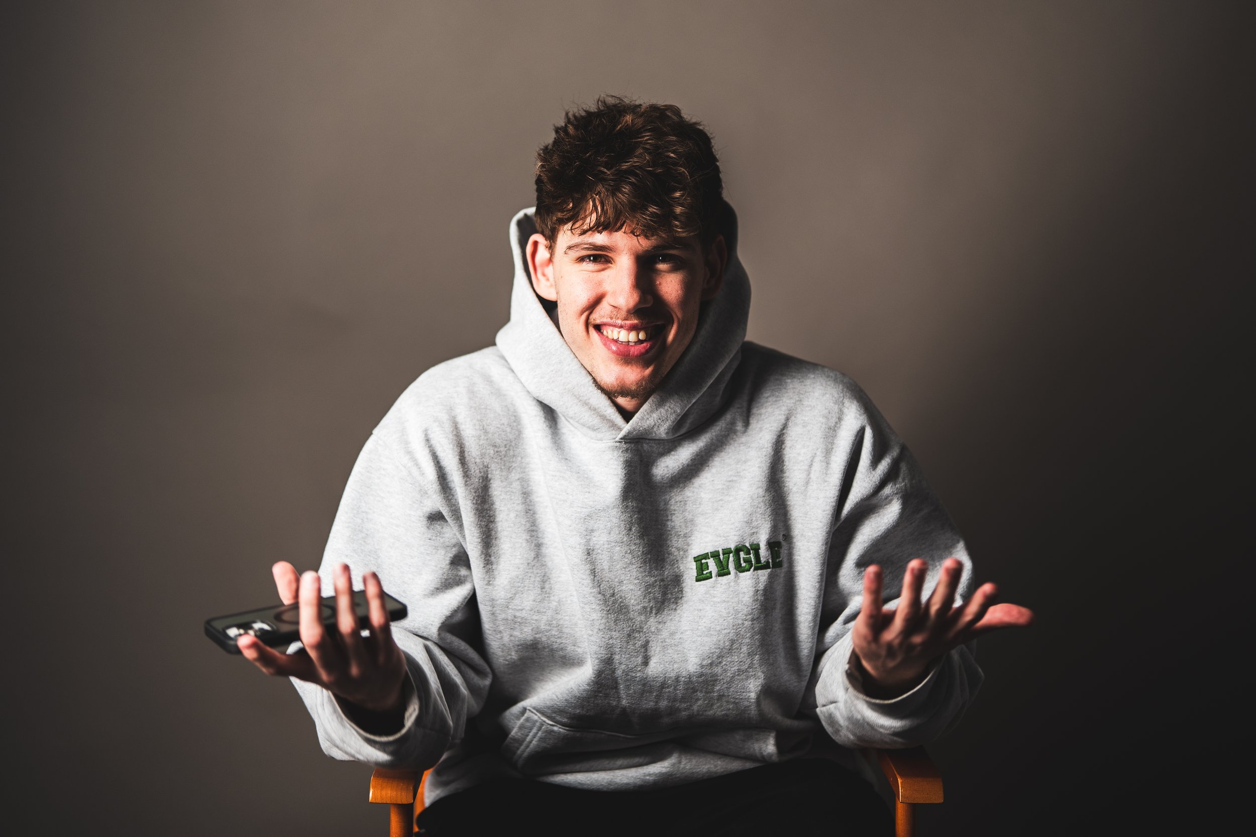 Young man sitting on a chair, wearing a gray hoodie with 'EVGL' logo, smiling, holding a phone, against a dark gray background.