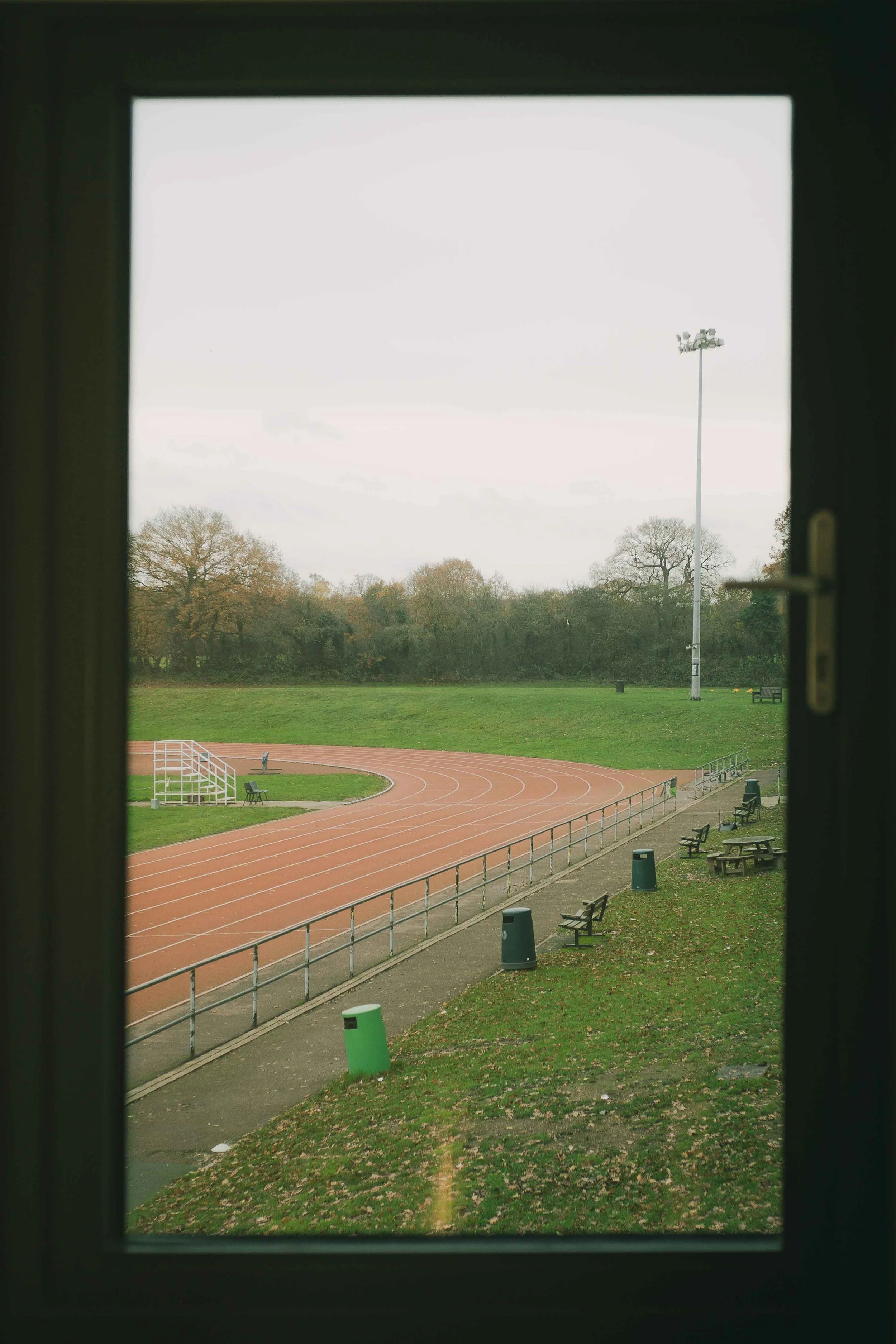 View of an outdoor running track seen through a window, with benches, trash cans, and trees in the background on a cloudy day.