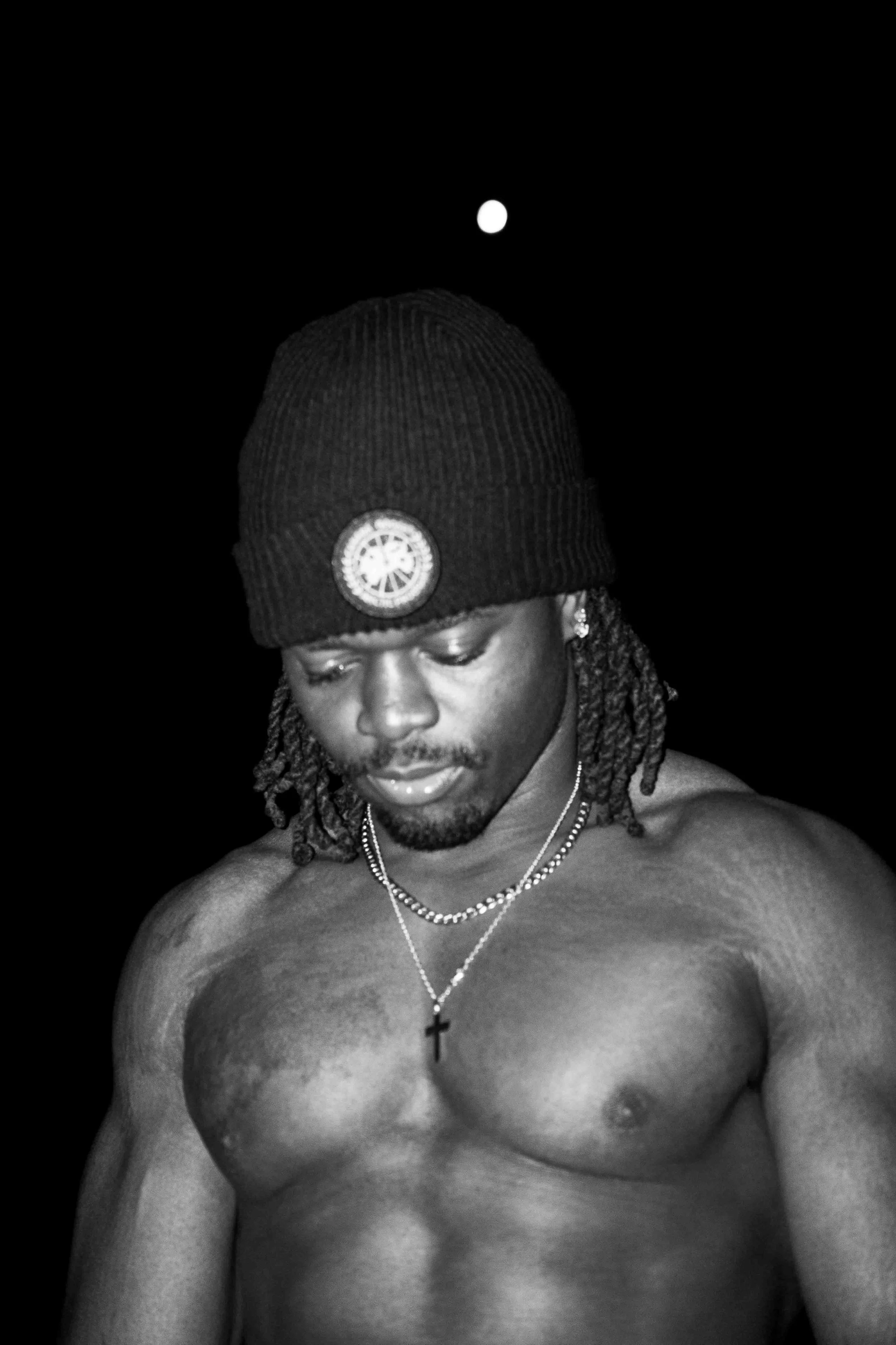 A shirtless man with dreadlocks wearing a black knit cap and a cross necklace, looking down, against a black background with a small white moon visible.