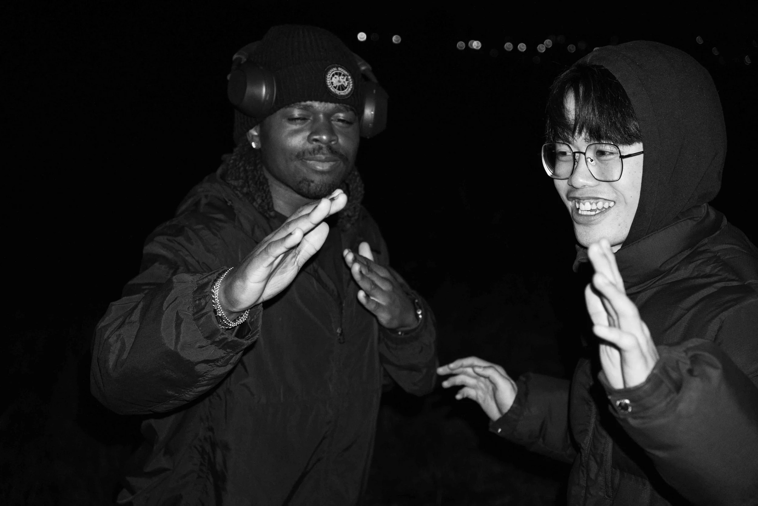 Two young men smiling and making hand gestures in a nighttime outdoor setting, with one wearing headphones and a beanie, and the other wearing glasses and a hooded jacket.