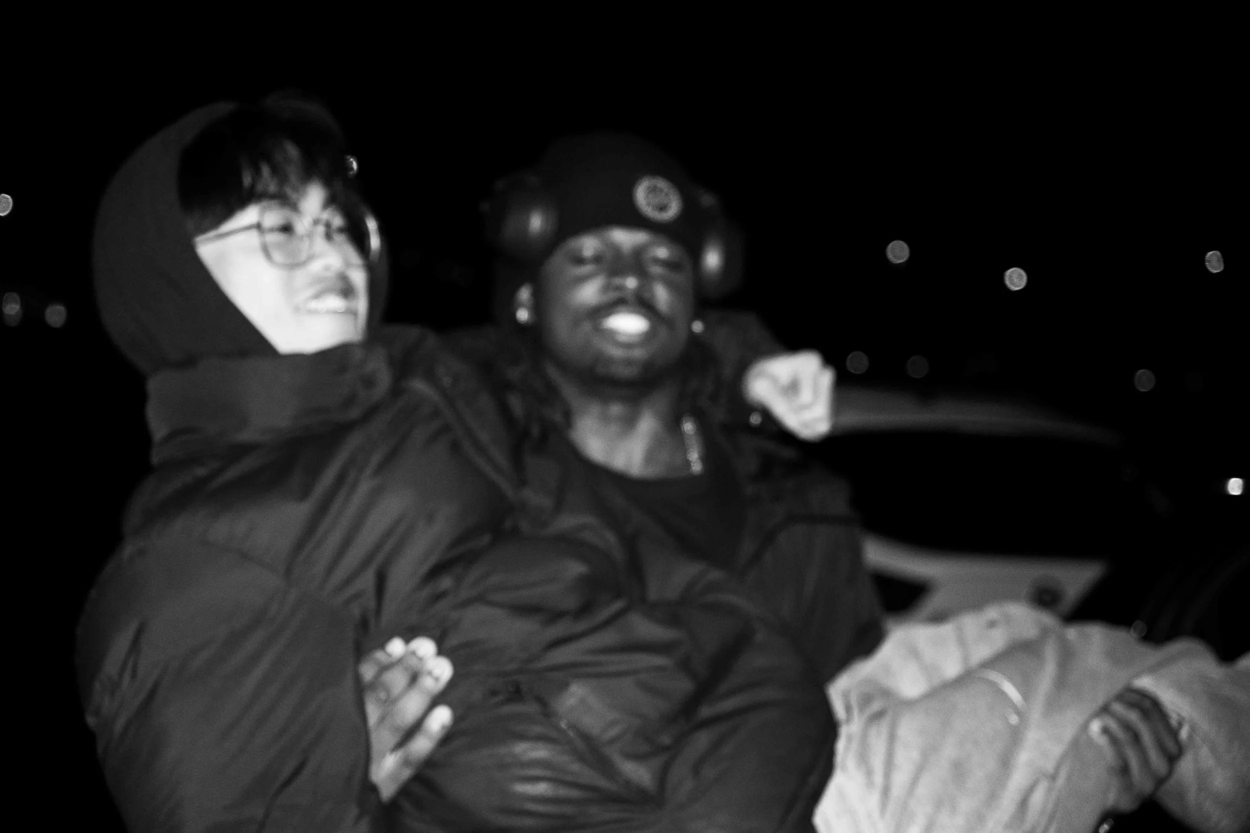 Two people sitting together at night, one wearing glasses and a hooded jacket smiling, the other wearing a hat with headphones and a jacket, smiling.