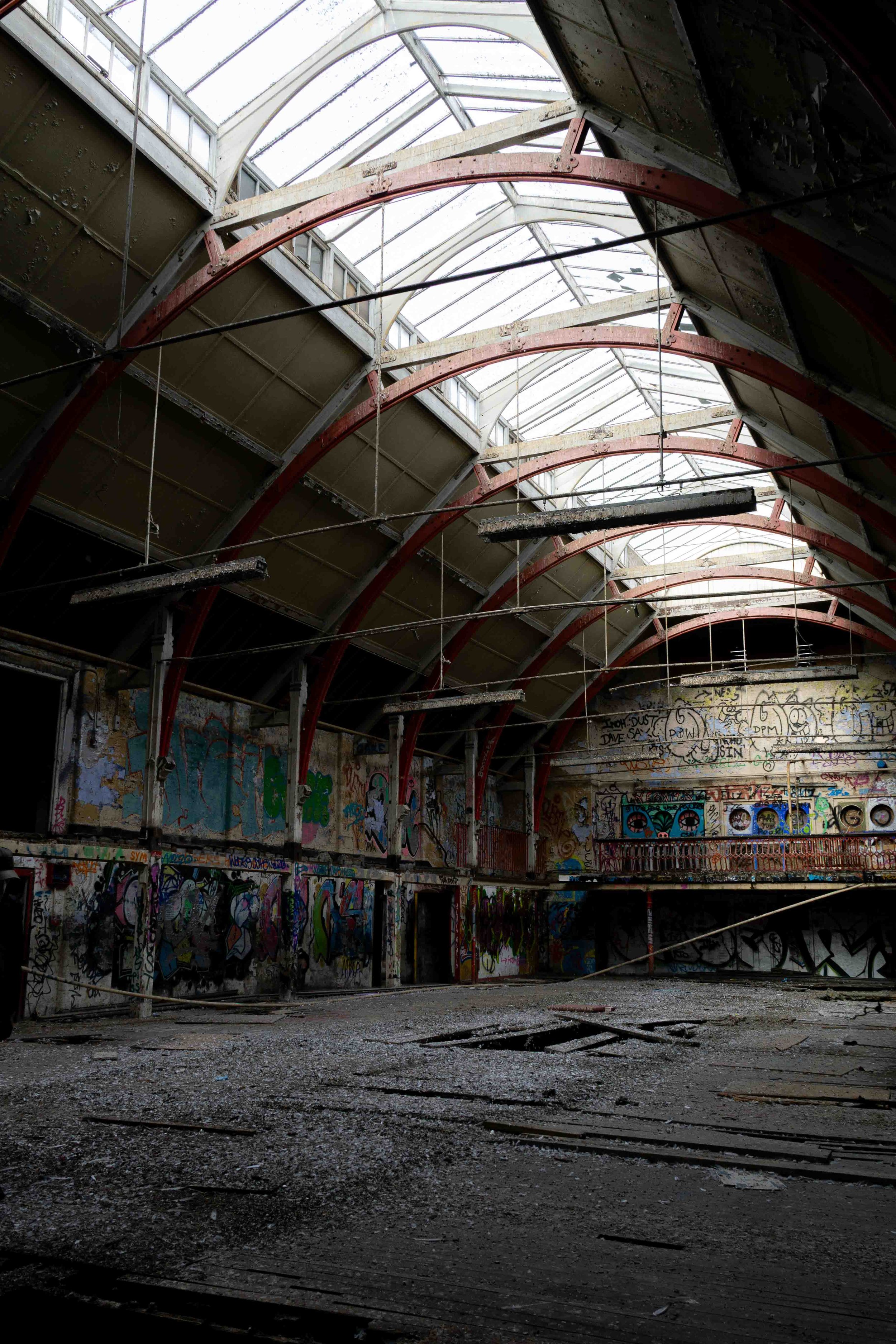 Interior of an abandoned building with graffiti-covered walls, a damaged floor, and a semi-arched glass ceiling.