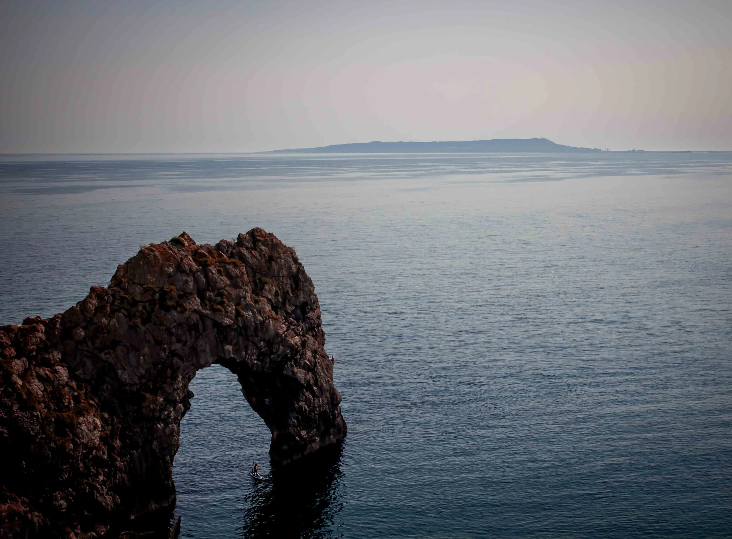 A large rock formation with a natural arch in the ocean, with a small boat near the arch and an island visible in the distance under a foggy sky.