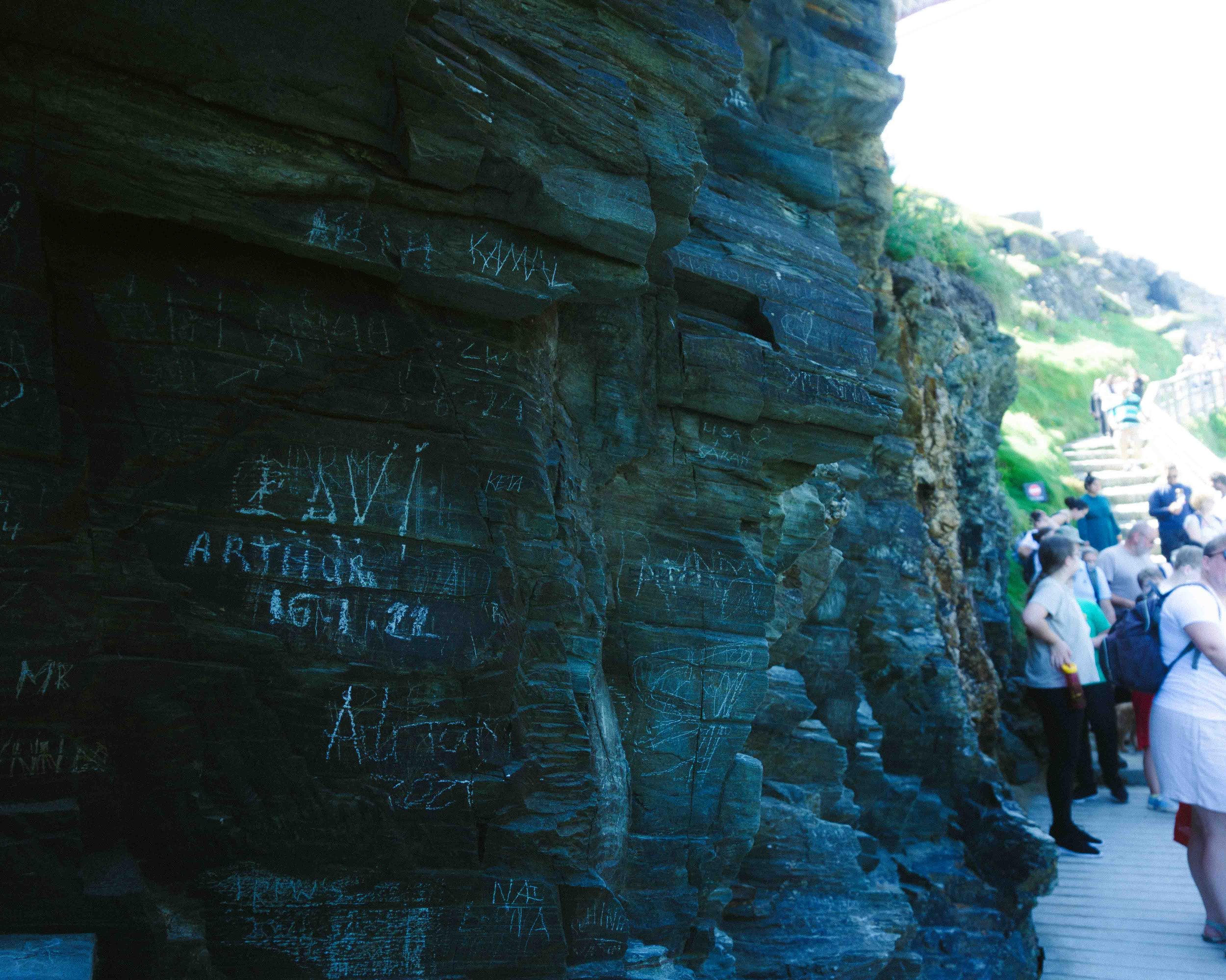 Rock formation with chalk graffiti on the left side and a crowd of people walking along a meandering wooden pathway on the right side, in a scenic outdoor location.