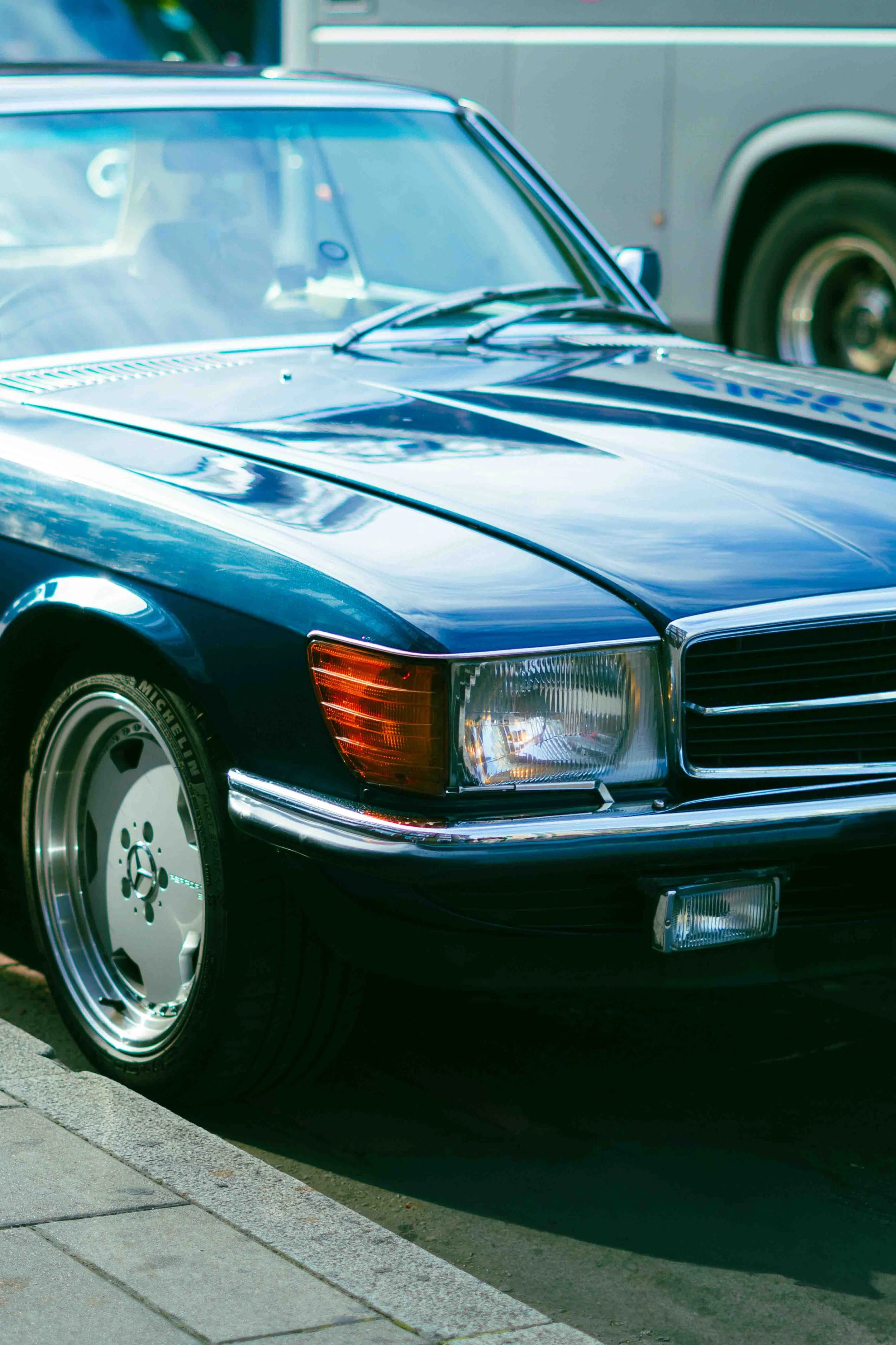 A close-up of the front of a vintage dark blue Mercedes-Benz car parked on a street, with another vehicle visible in the background.
