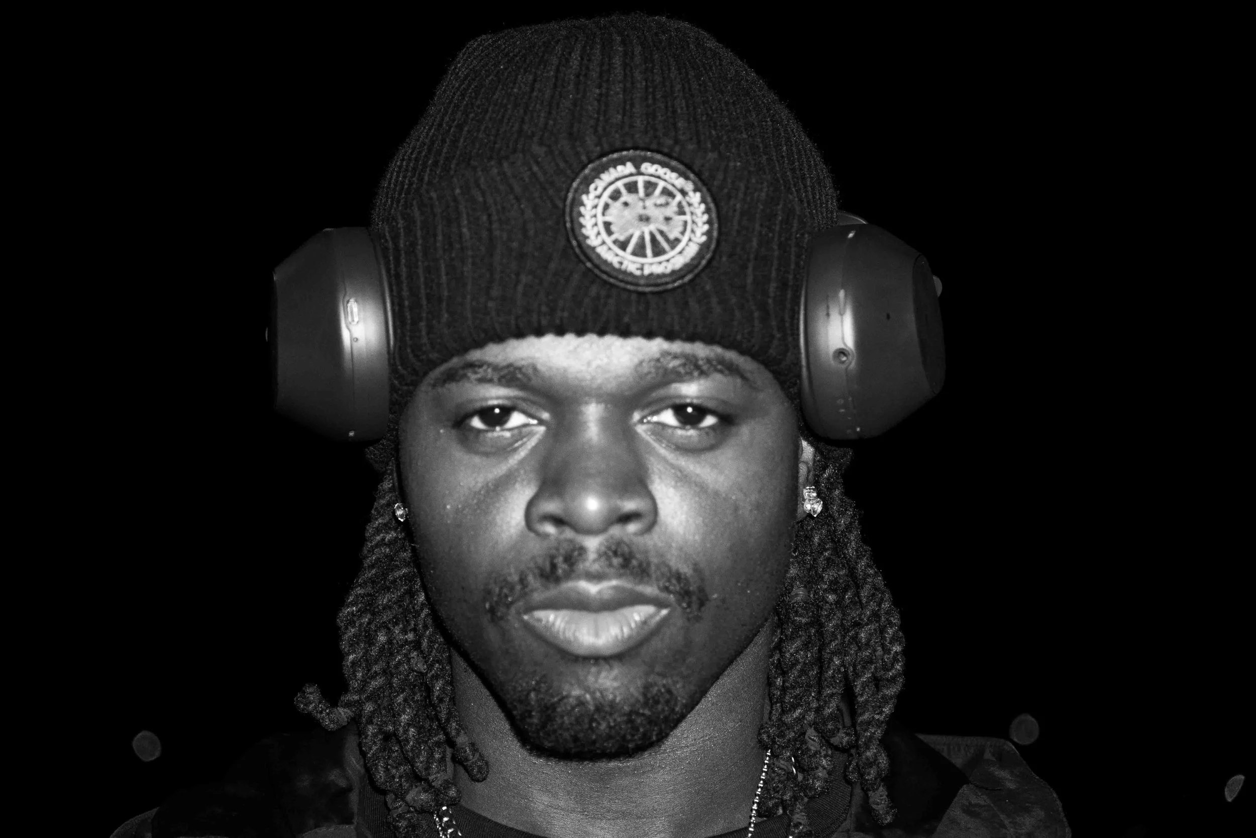 A young man wearing a beanie with a logo, headphones, and earrings, looking directly at the camera against a black background.