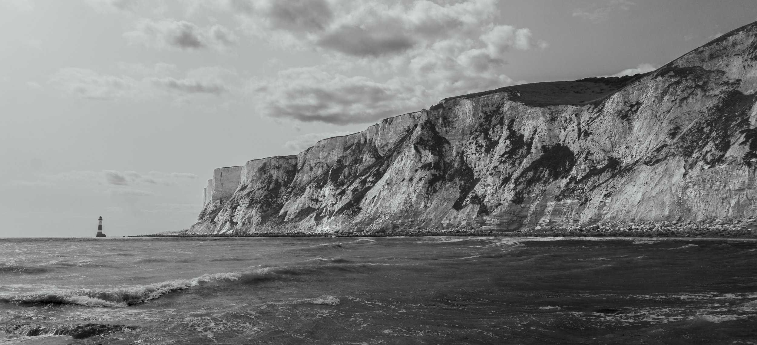 Black and white photograph of a rugged coastline with steep cliffs and a lighthouse in the distance on the water. The ocean has waves, and the sky has scattered clouds.