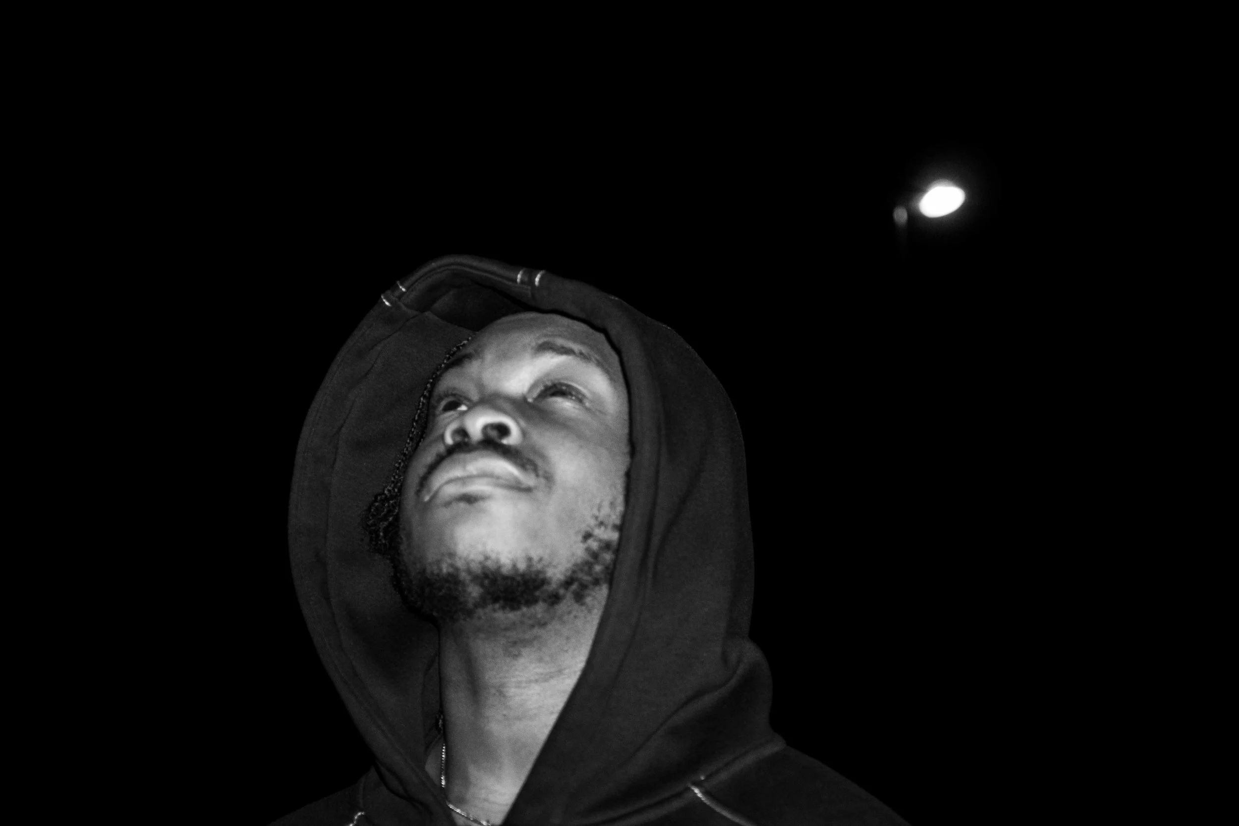 A man in a black hoodie looking upward against a dark night sky with a bright moon.