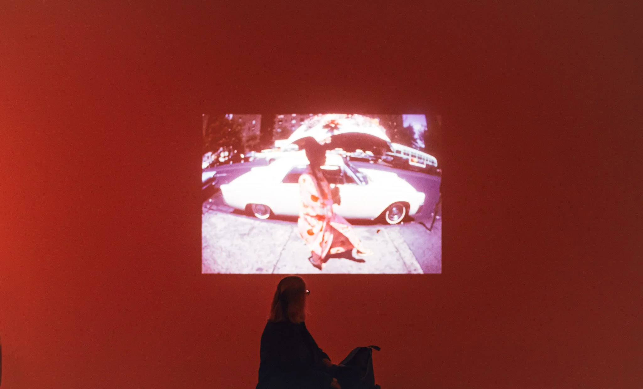 A person sitting and looking at a projected image on a red wall. The projected image shows a woman in a traditional Japanese kimono walking in front of a vintage white car, with trees and buildings in the background.