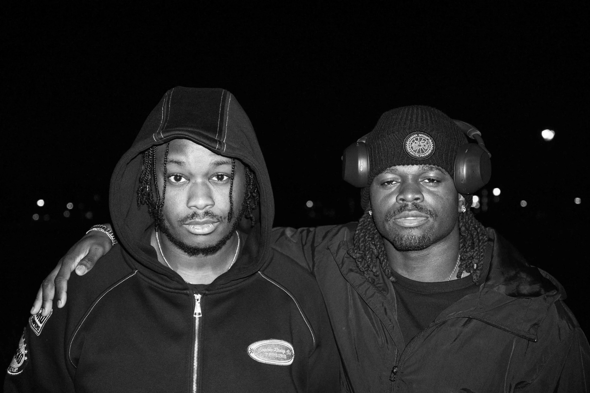 Two young men standing close together, one with his arm around the other's shoulder, in a nighttime outdoor setting. The man on the left is wearing a hooded sweatshirt with the hood up, and the man on the right is wearing a beanie hat with headphones