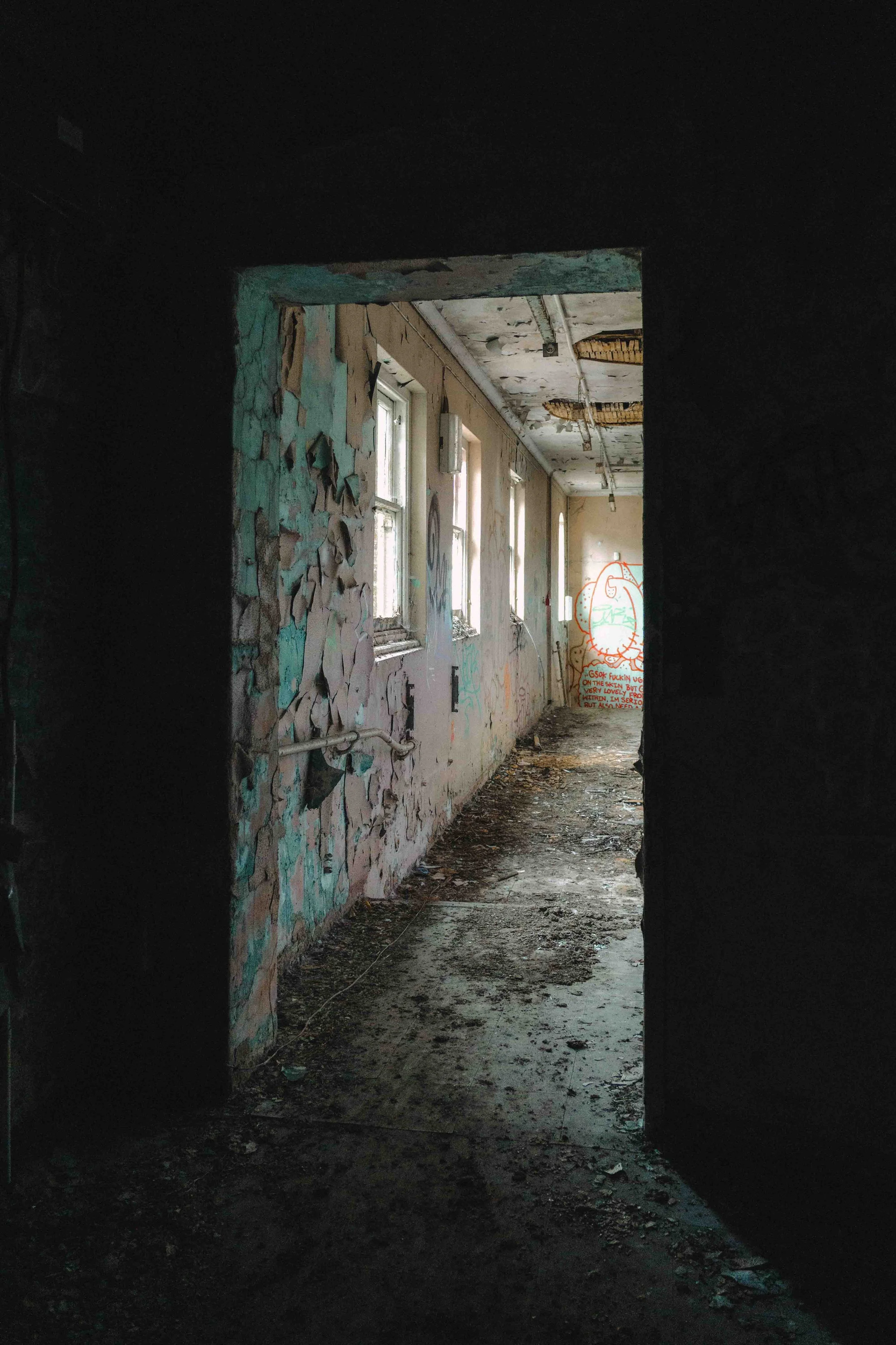 View through an arched doorway into an abandoned building with peeling paint, graffiti on the walls, broken ceiling panels, and a dirt-covered floor. Graffiti on the back wall includes a shape resembling a face and some text.