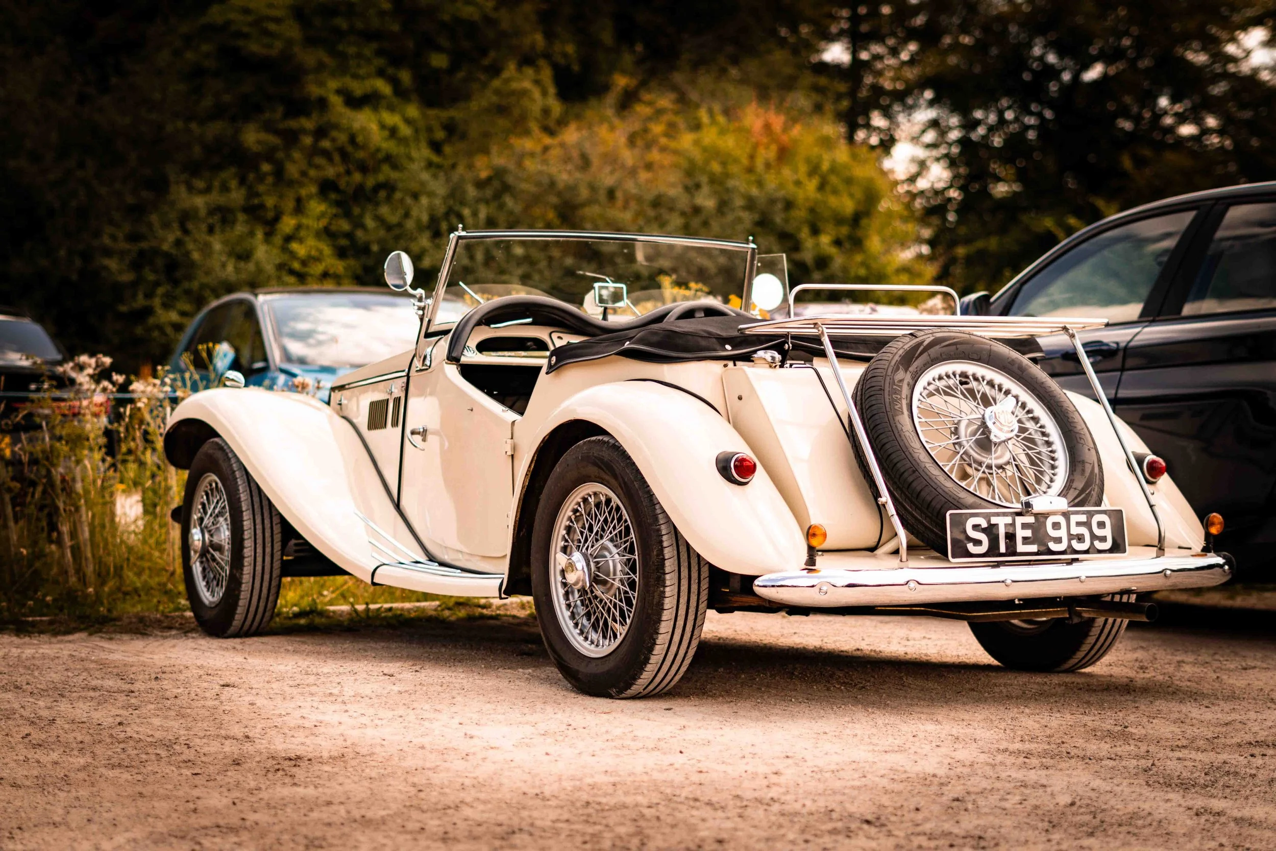 Vintage white convertible car with a spare tire mounted on the back, parked on a dirt surface with trees in the background.