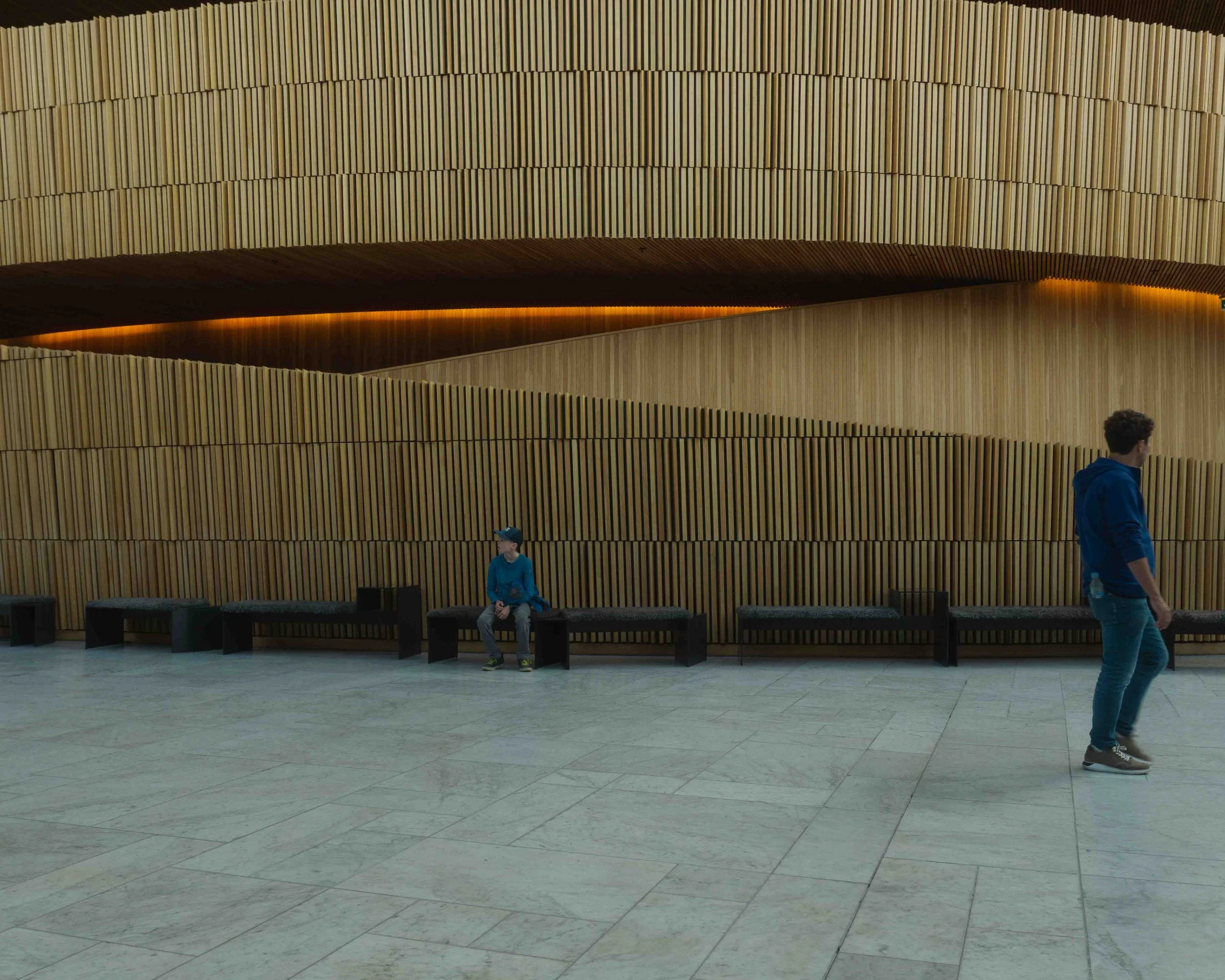 Interior of a modern building with wooden panel walls and black benches. Two people, a young boy sitting and an adult walking, are present in front of the wall.