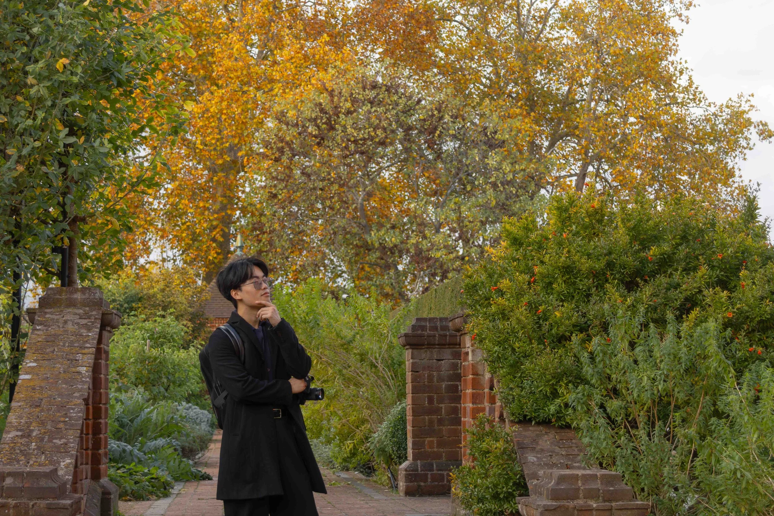 A young man with black hair and glasses stands on a brick pathway, surrounded by green bushes and trees with autumn-colored leaves. He is wearing a black coat, black pants, and has a small backpack. He is holding a camera in his left hand and appears to be pondering or observing the scene around him.