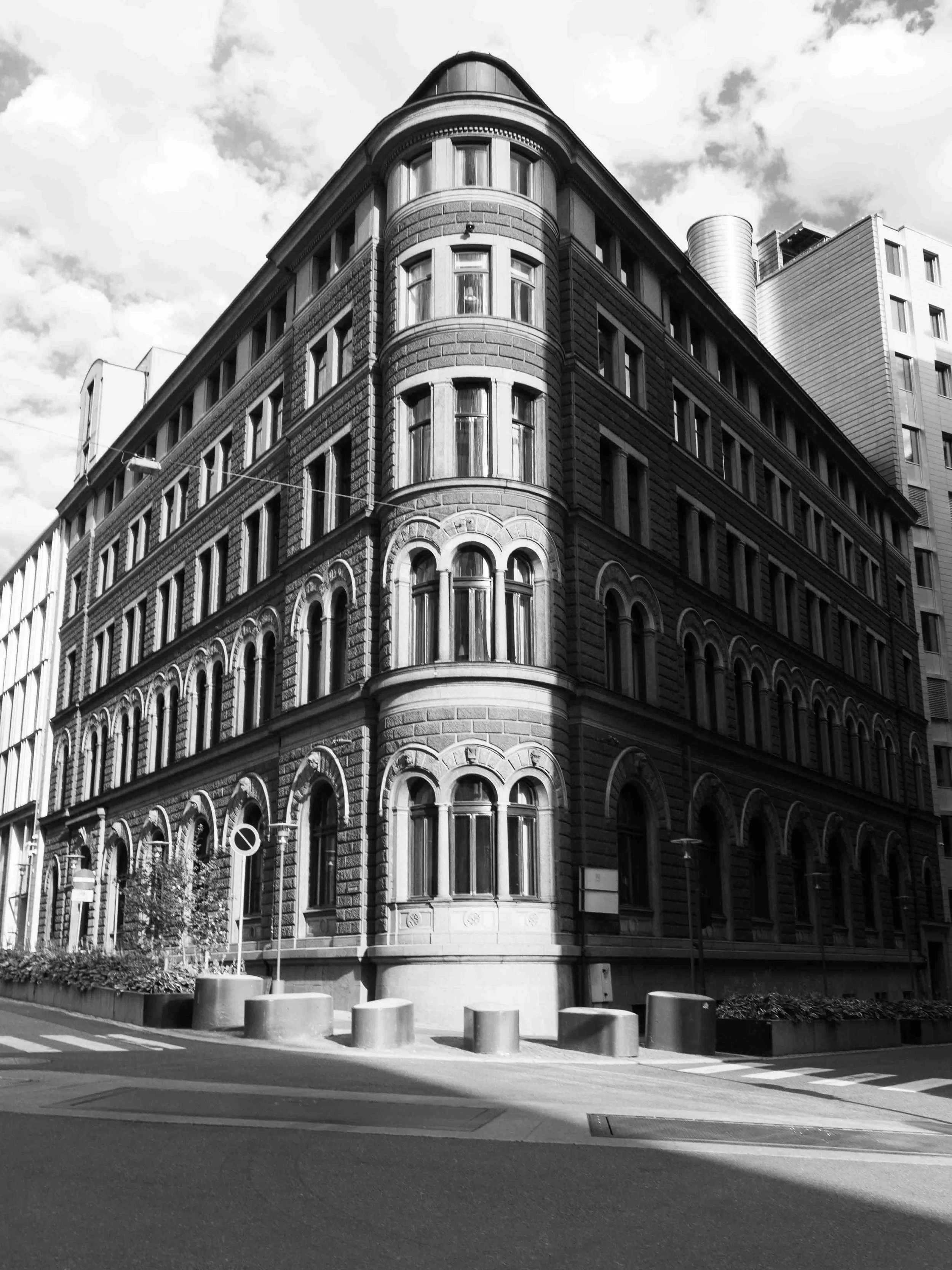 Black and white photo of a historic multi-story building with rounded corner architecture, ornate window details, and a modern building in the background.