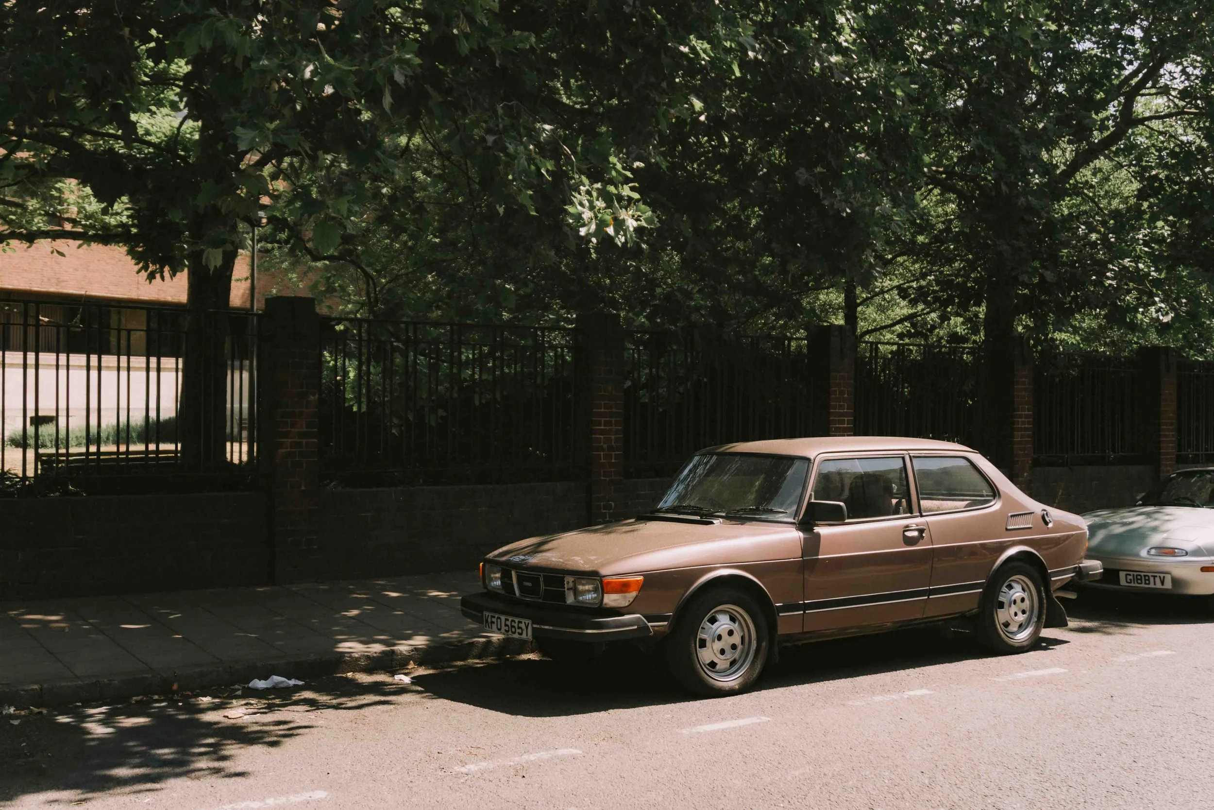 A vintage brown car parked on the street under green leafy trees. Another car is partially visible behind it.