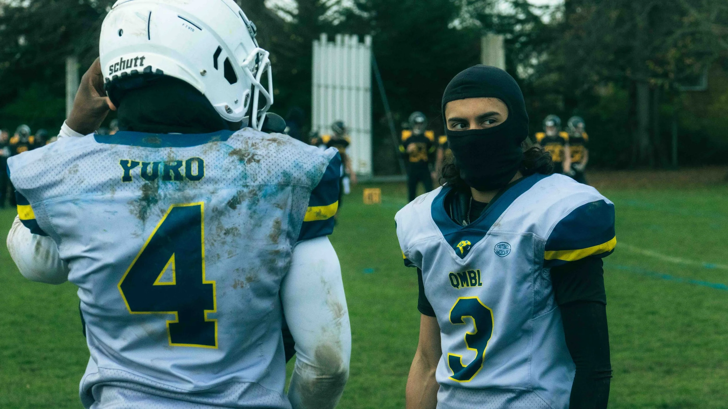 Two football players in helmets and jerseys on a field with a group of players in the background. One player is adjusting his helmet, and the other is wearing a facemask covering his nose and mouth, with long hair visible.