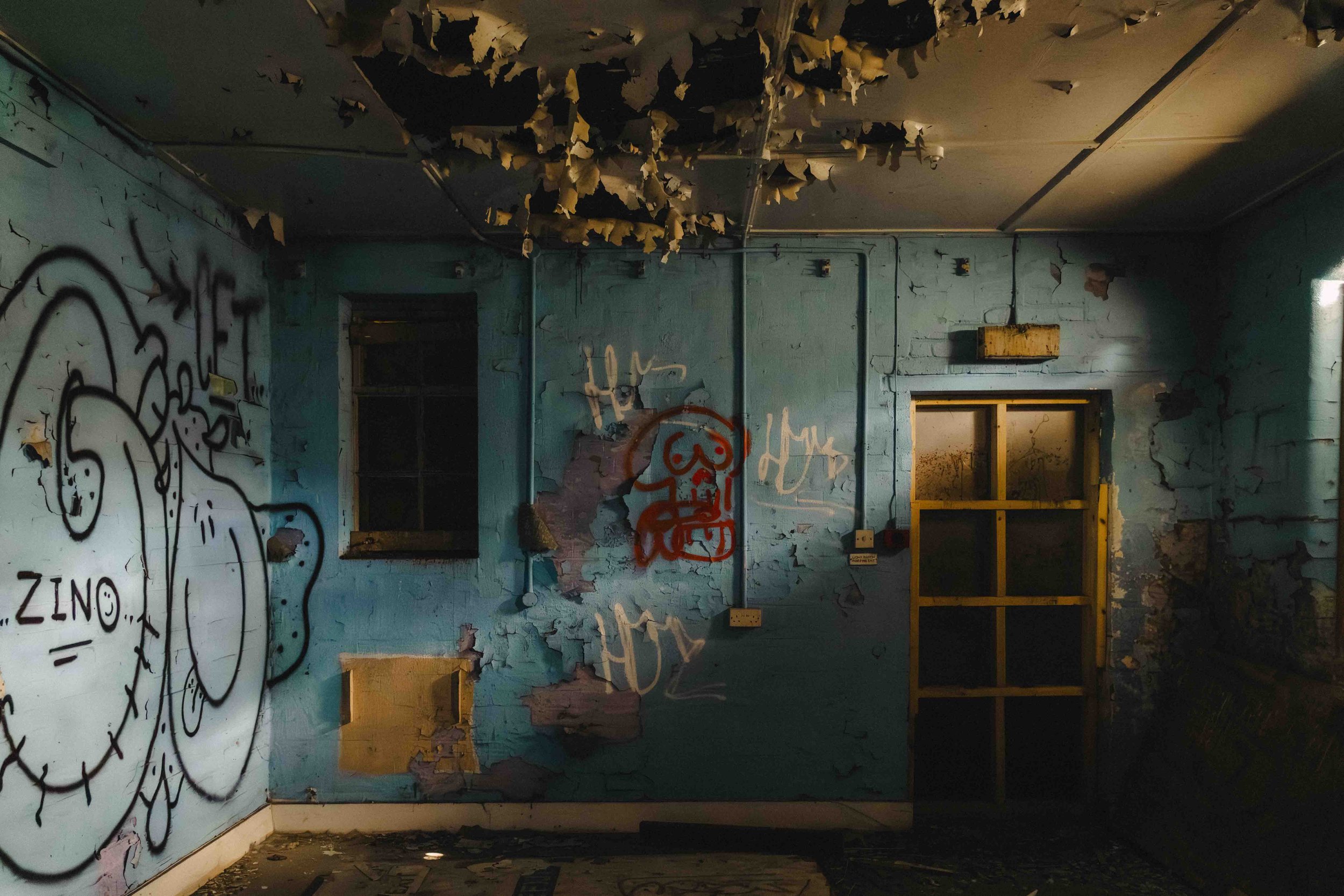An abandoned room with peeling blue paint, graffiti on the walls, broken ceiling tiles, a small yellow door frame, and a window with dirt and debris on the glass.