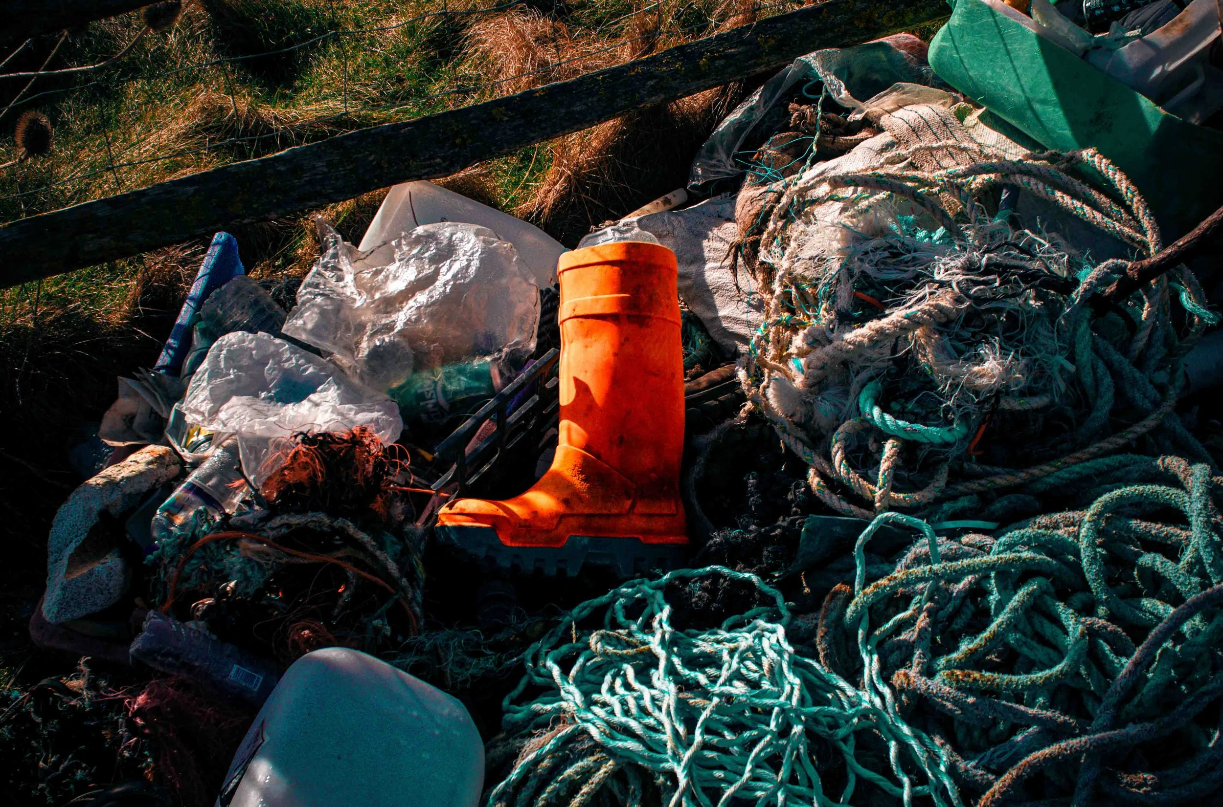 Pile of assorted debris including tangled ropes, a bright orange rubber boot, plastic bottles, and miscellaneous trash outdoors.