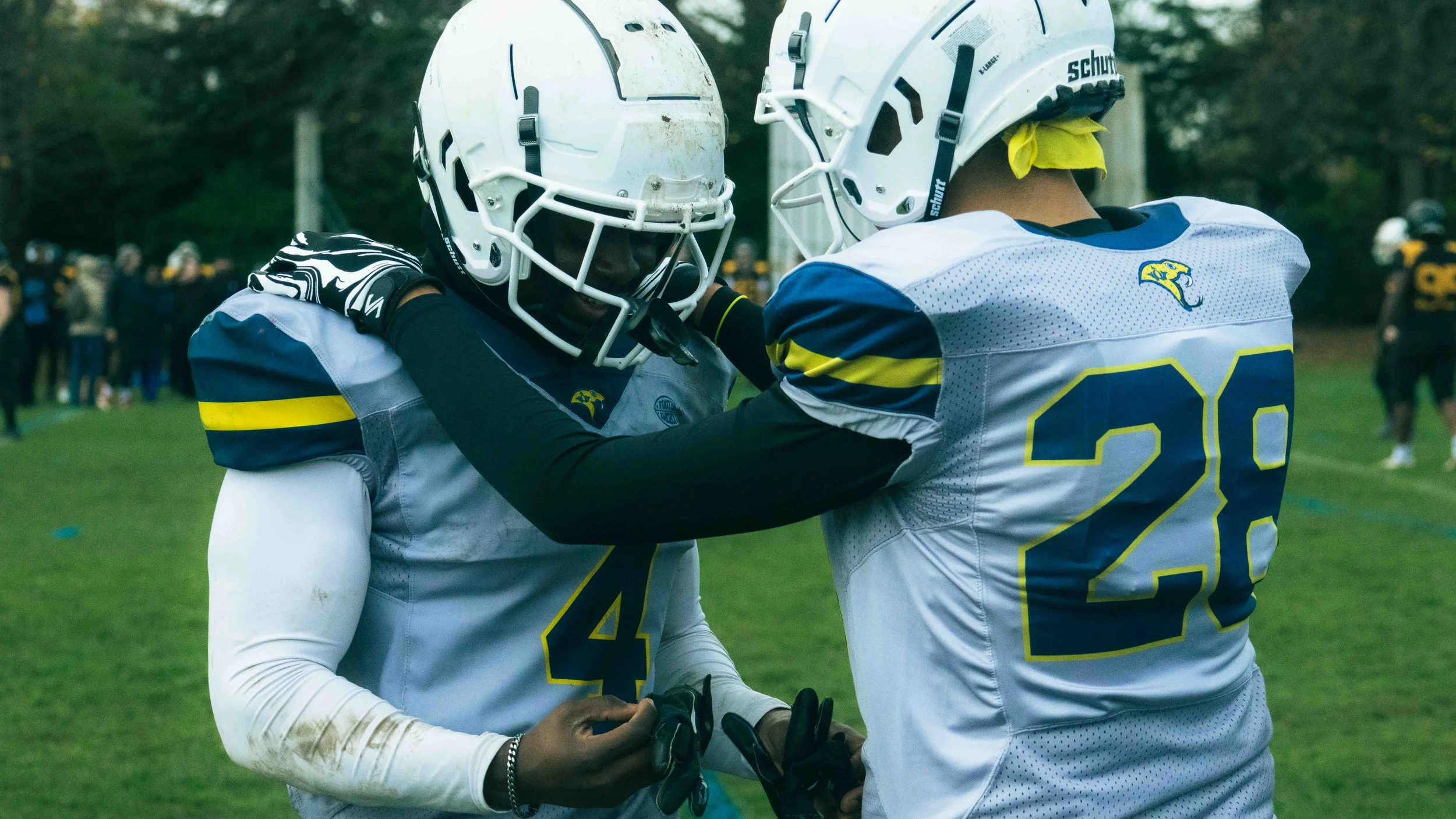 Two football players in white and blue uniforms, wearing helmets, embrace on a grassy field during a game, with spectators in the background.
