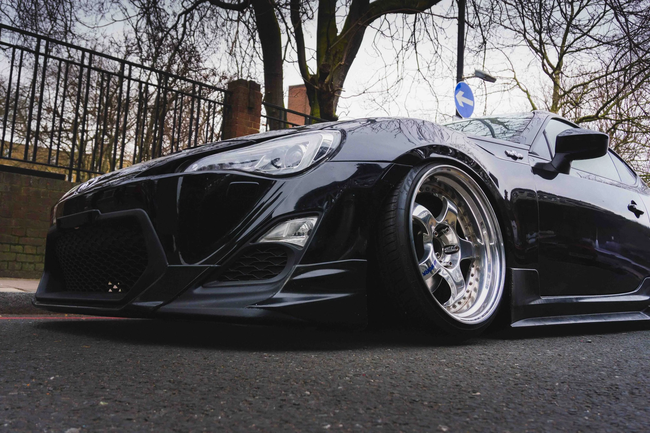 A black sports car parked on the street with a low stance and custom wheels, background of trees and a fence.