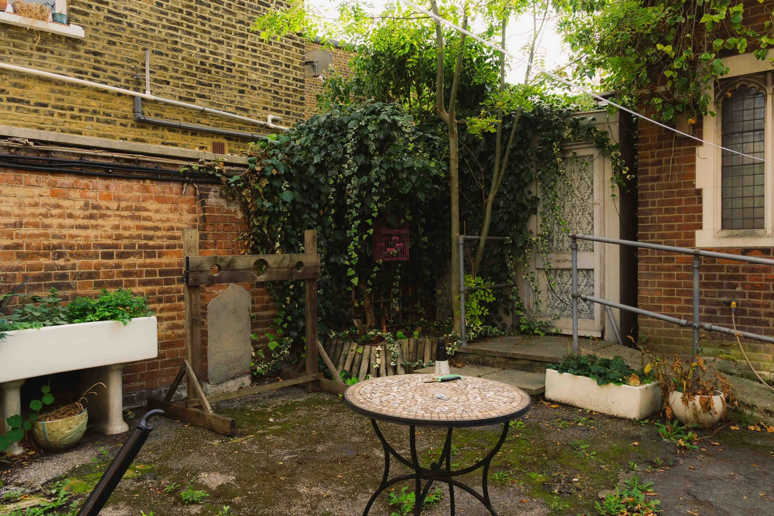 A small urban backyard patio with a round mosaic table, potted plants, and overgrown greenery, surrounded by brick walls and an exterior door with lace curtains.