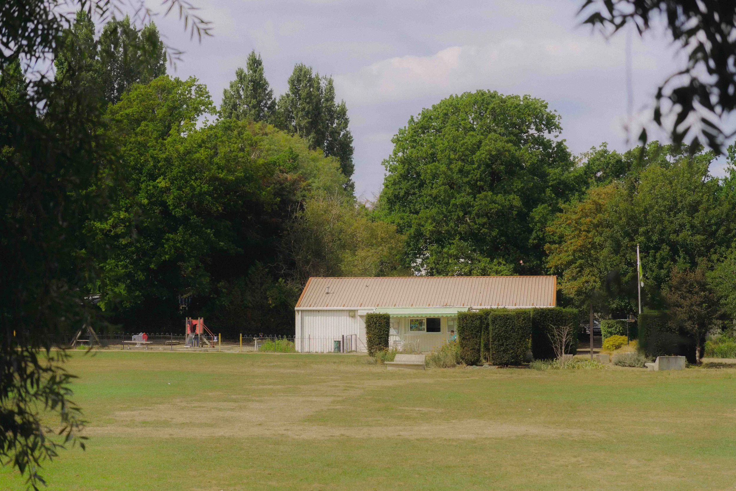 A park with a large grassy area, surrounded by trees, a small white building, and a playground in the background.