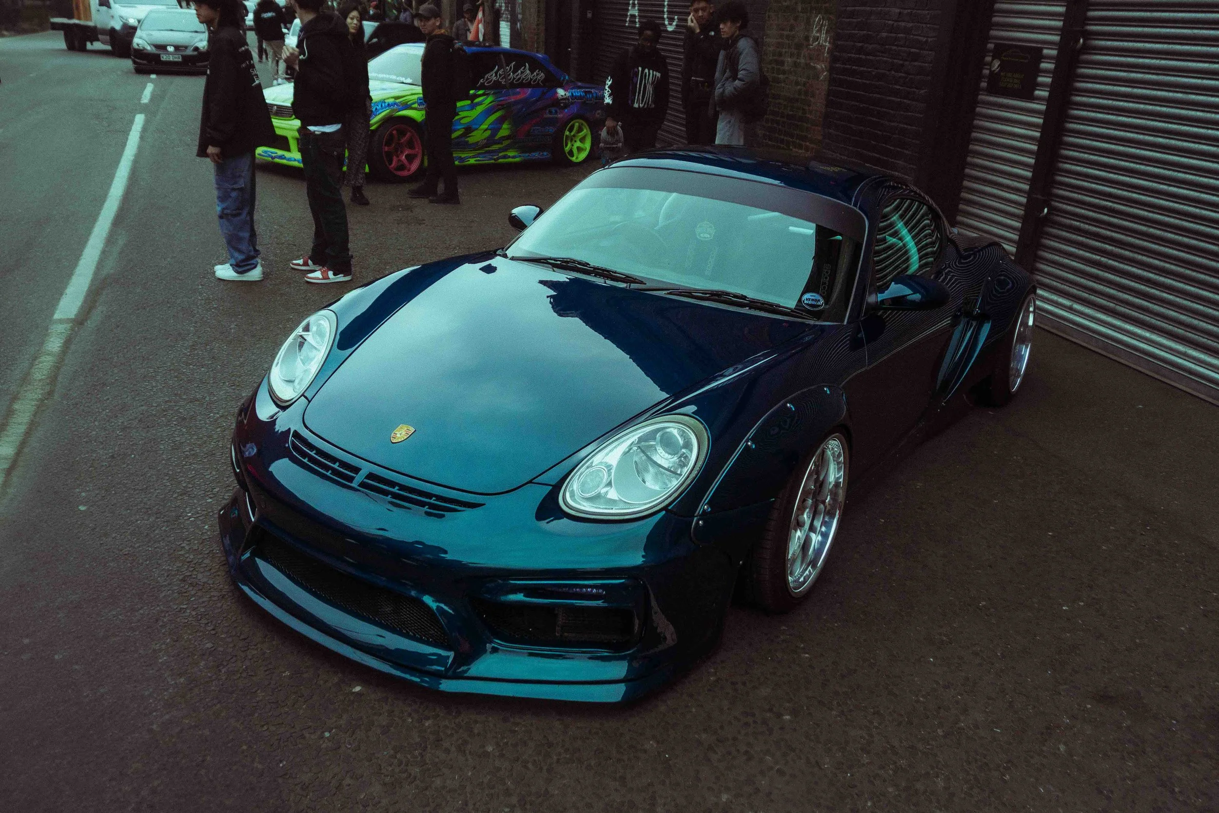 A dark blue Porsche sports car parked on a street in front of a brick building with a rolling shutter. Several people stand nearby, and a colorful car is visible in the background.