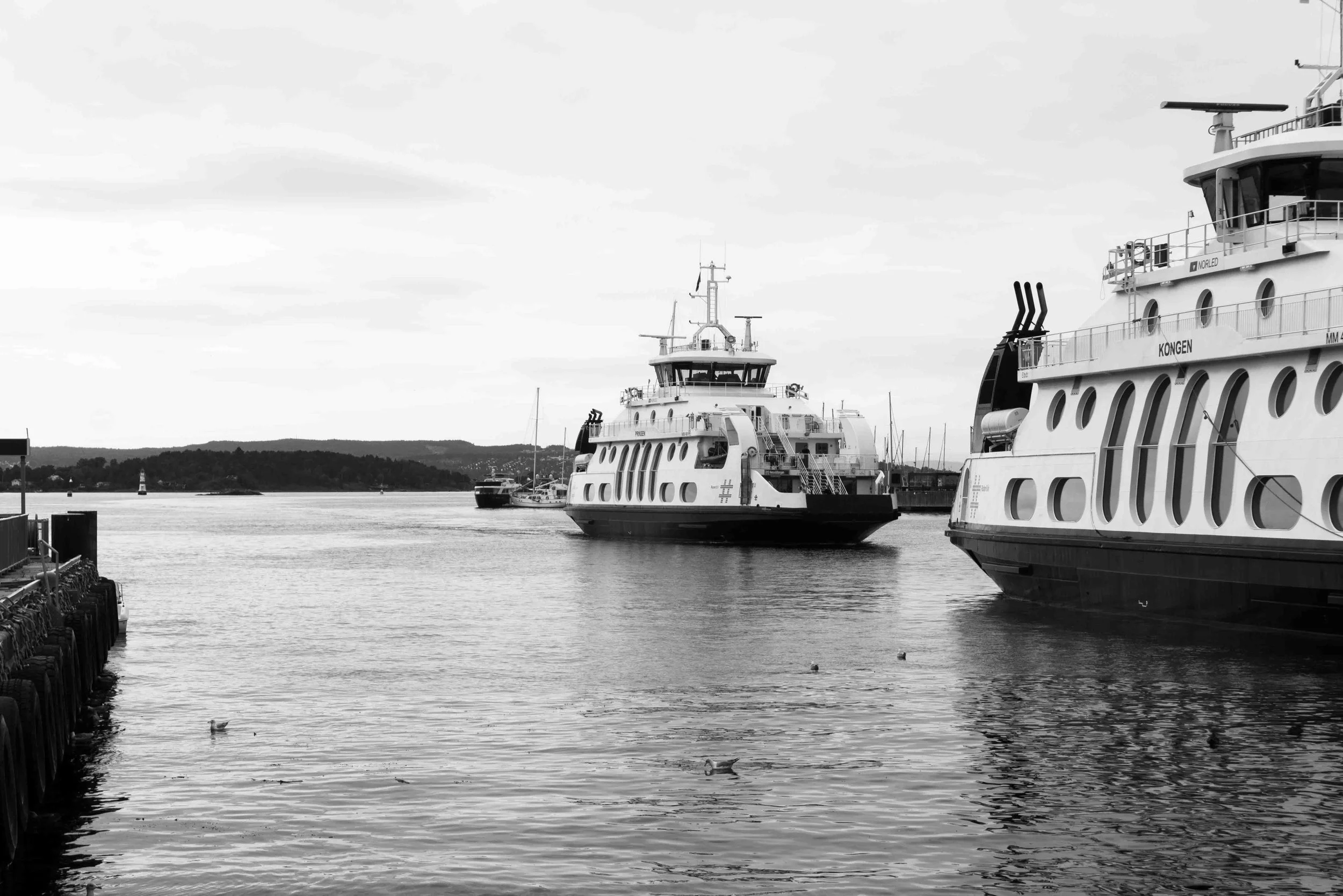 Black and white photo of three docked boats or ferries in a harbor with a distant shoreline and cloudy sky.