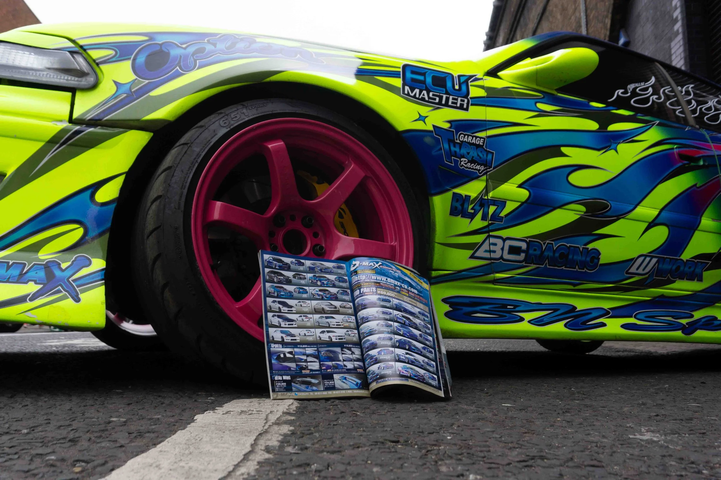 Close-up of a brightly colored race car with pink wheels, blue and yellow flame decals, and various sponsor logos, parked on an asphalt surface with a brochure or magazine open on the ground in front of it.