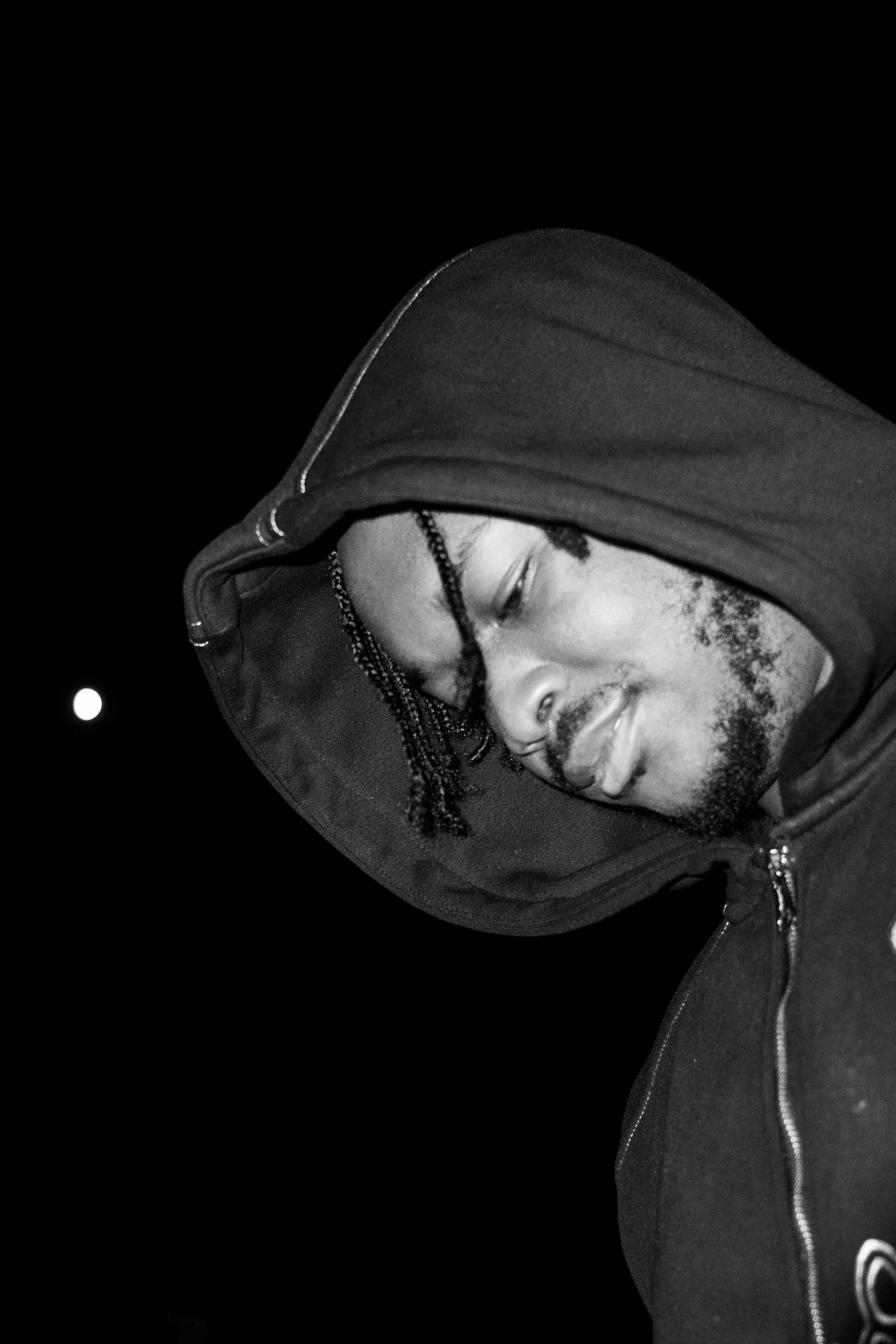 A young man with braided hair, a beard, and closed eyes wearing a hooded jacket against a dark background with a small bright light