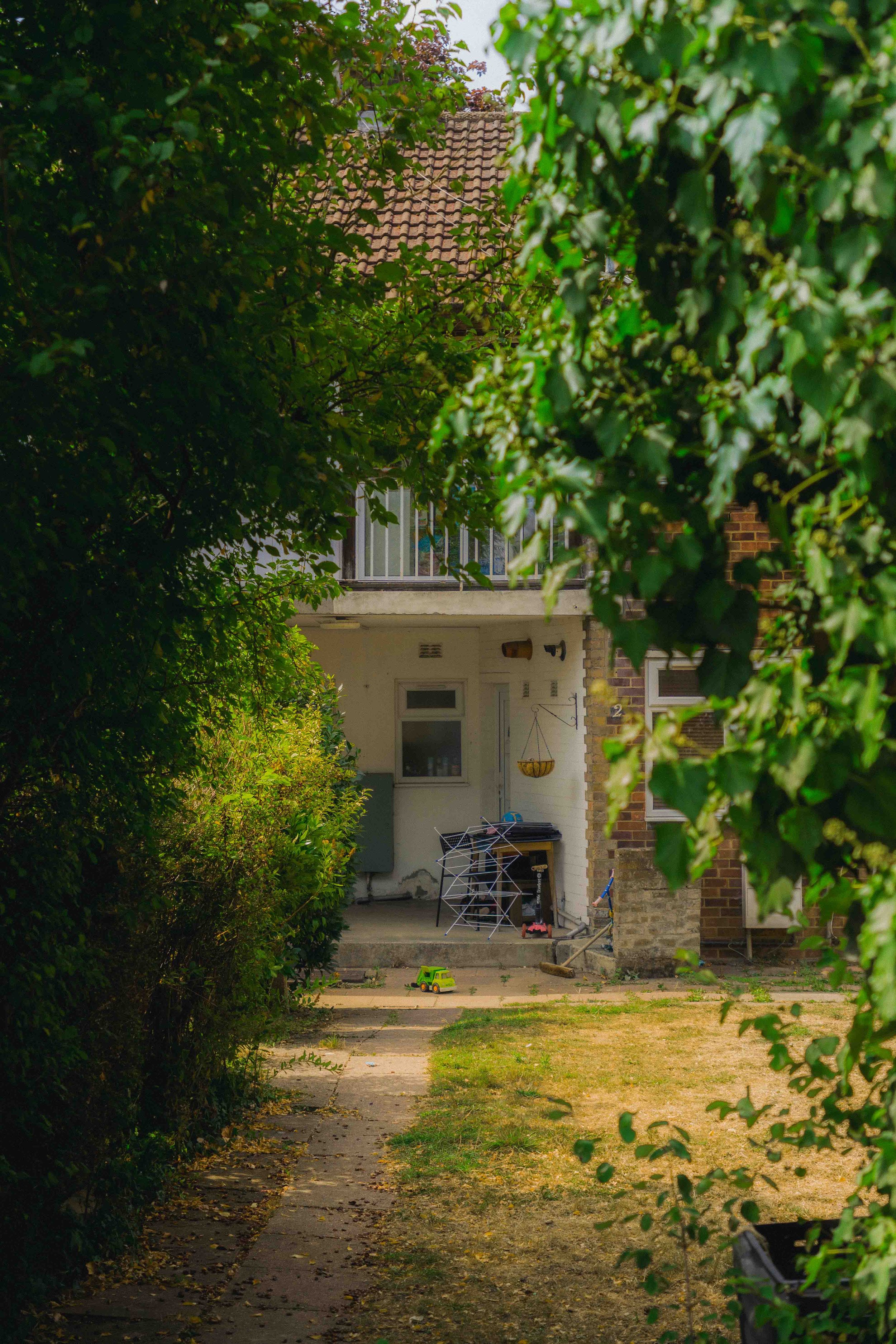 View of a backyard with a pathway leading to a house entrance, surrounded by greenery and bushes on both sides, with a small toy truck on the ground and various outdoor objects near the house entrance.