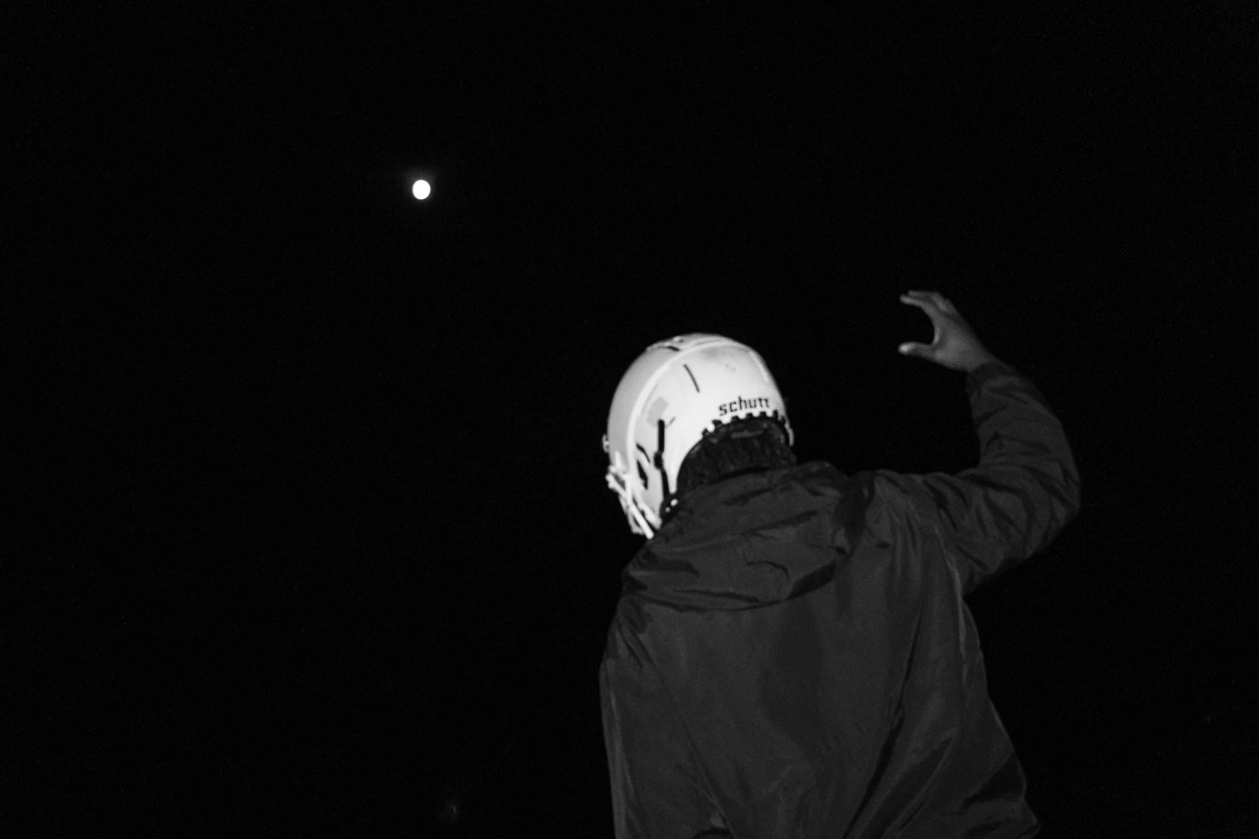 A person wearing a helmet and jacket reaching towards the night sky where a bright object, possibly the moon or a star, is visible.