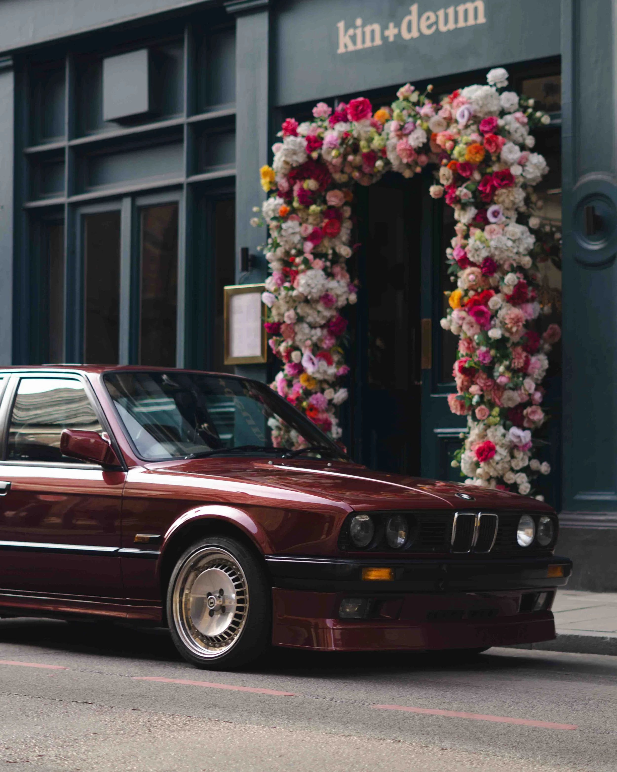 A vintage maroon BMW parked in front of a building with the sign 'kin+deum' and a large floral archway entrance decorated with pink, white, and yellow flowers.