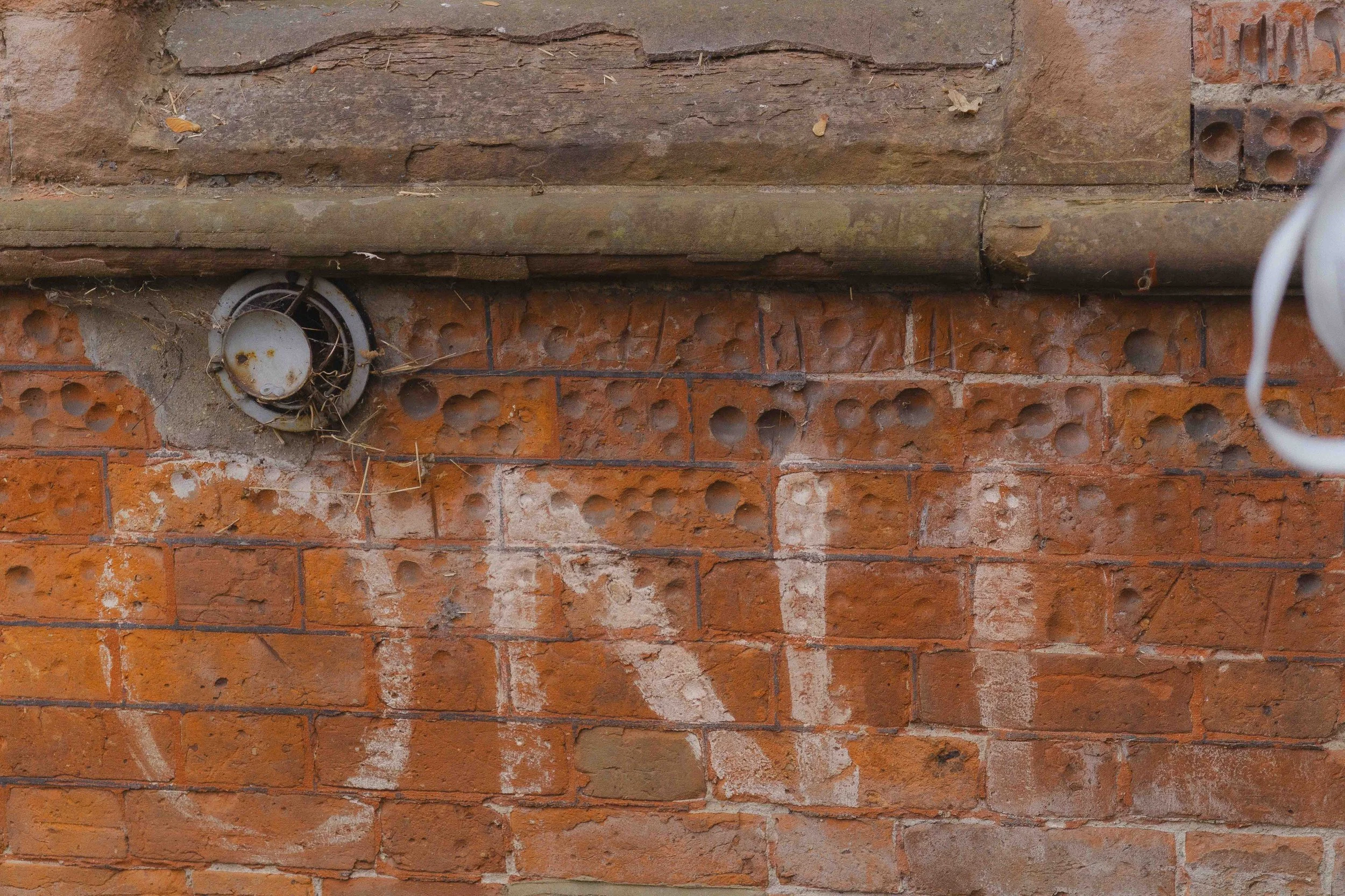 A brick wall with some graffiti, an air vent with debris on it, and a wooden ledge at the top.
