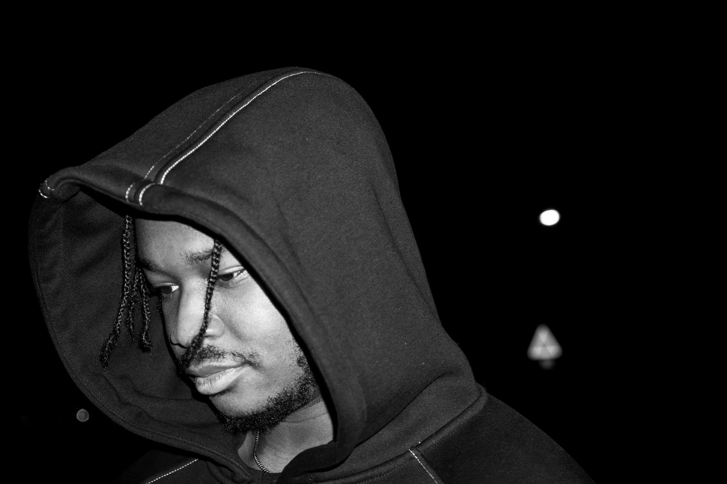 Close-up of a young man wearing a black hoodie at night, with a dark background and visible street signs.