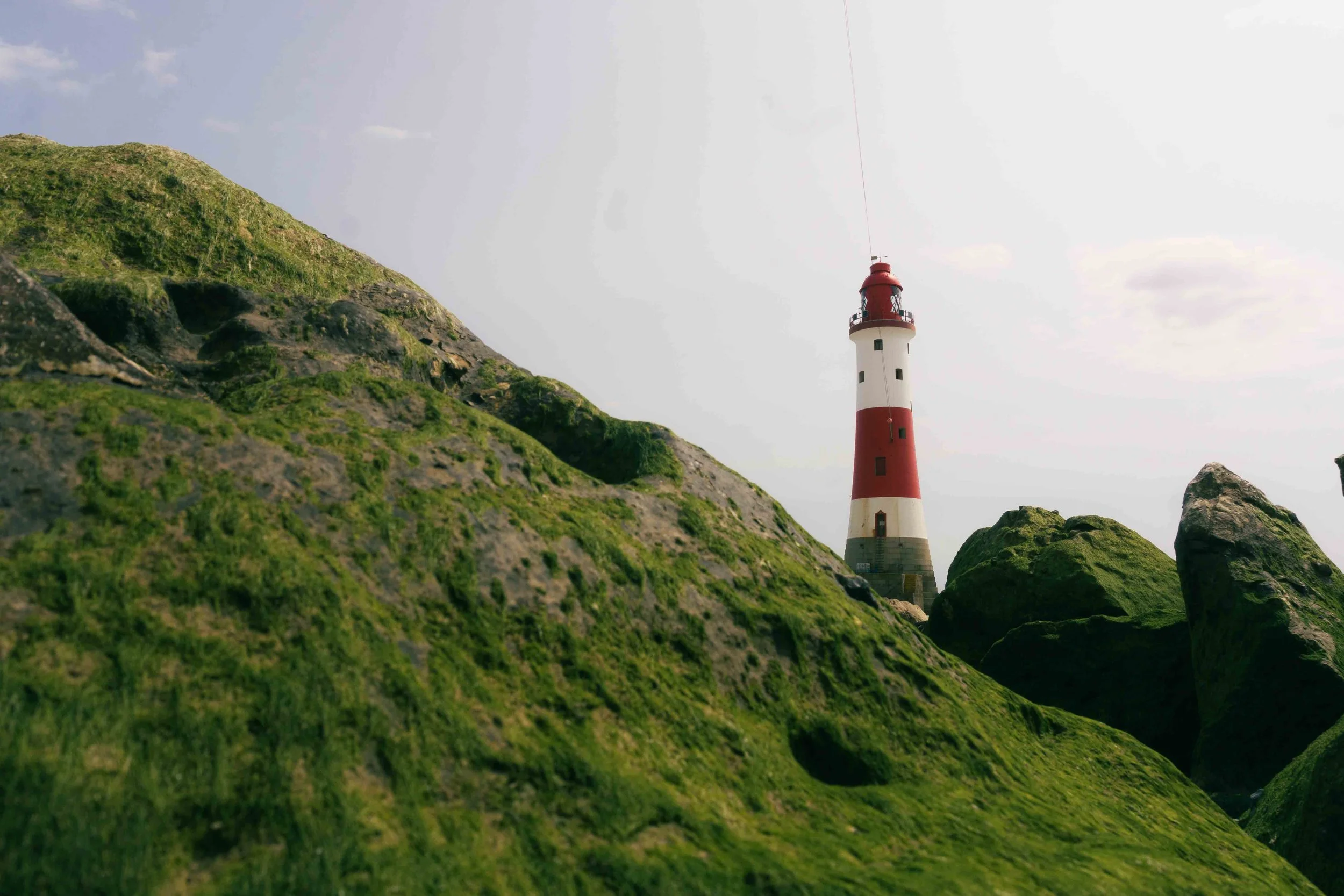 A lighthouse with red and white stripes standing among large moss-covered rocks and hills against a gray sky.