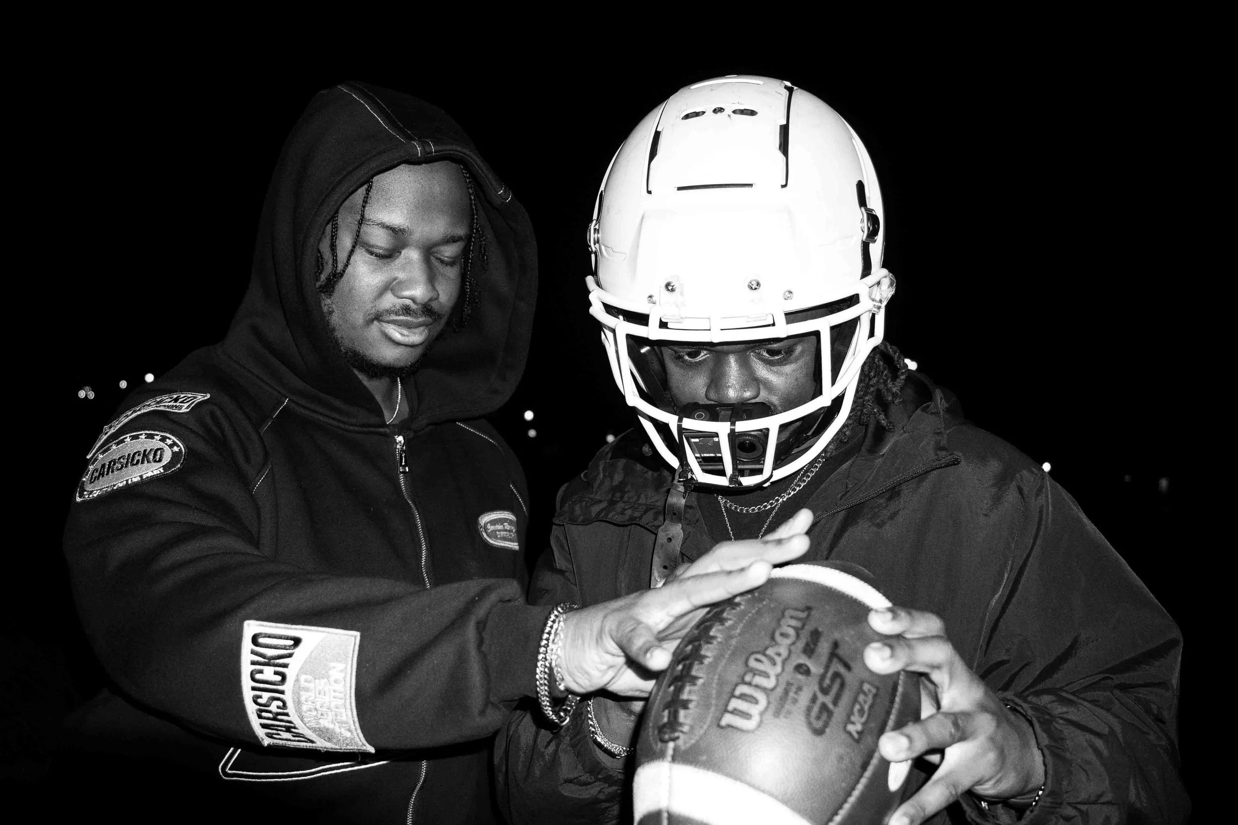 Two men, one wearing a football helmet and coat, and the other wearing a jacket with patches, are looking at a football together at night.