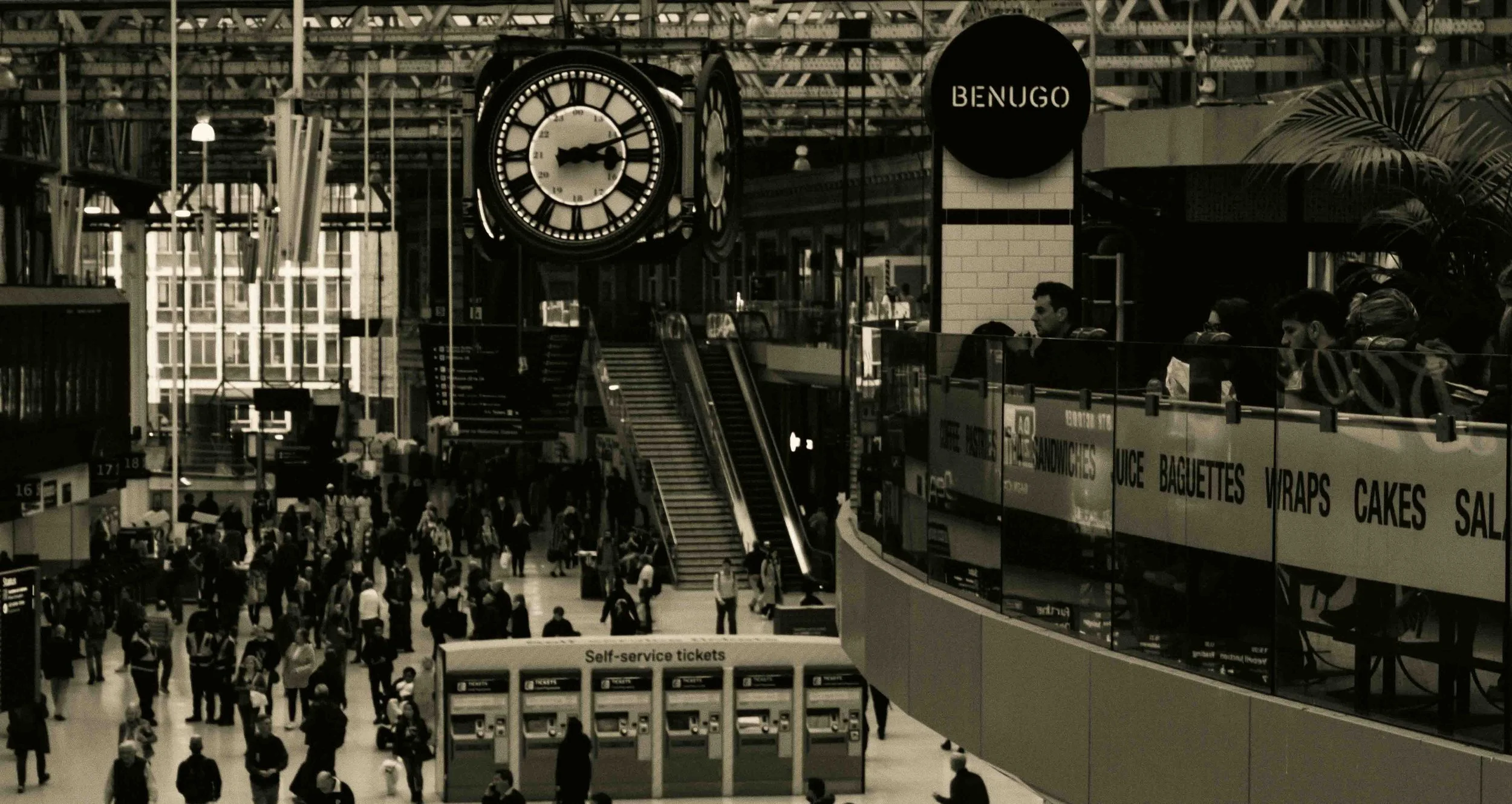 A busy train station concourse with a large clock showing approximately 3:12, people walking and standing around, an escalator leading upstairs, a sign reading 'BENUGO', and self-service ticket machines.
