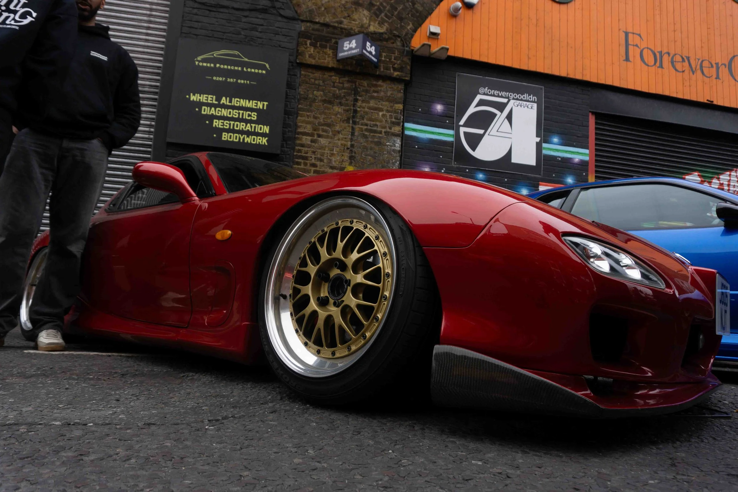 Red sports car with gold wheels parked on a city street, with part of a person visible on the left and buildings with signs in the background.