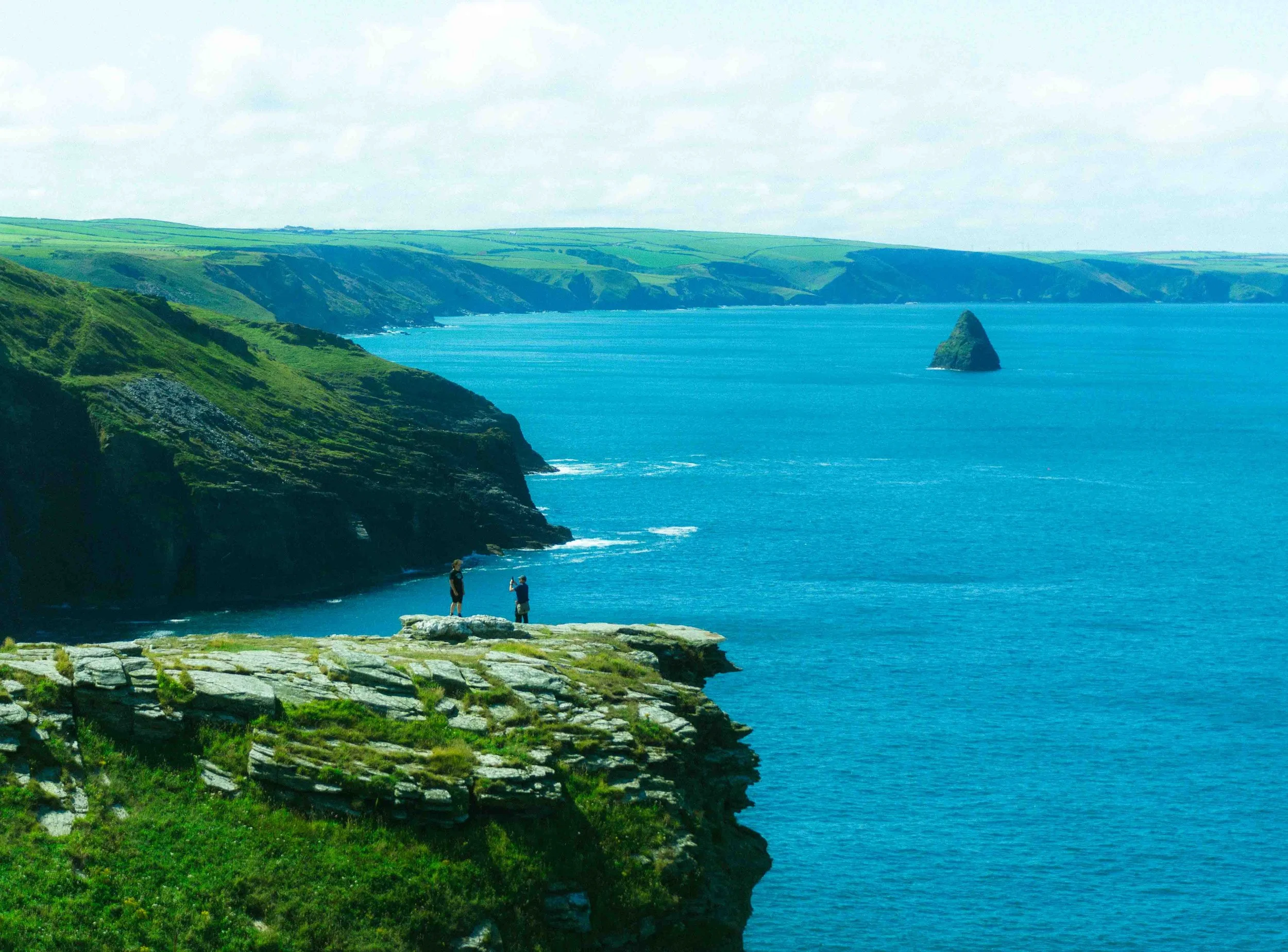 Two people standing on a rocky cliff overlooking a blue ocean with green hills and a large rock formation in the water.