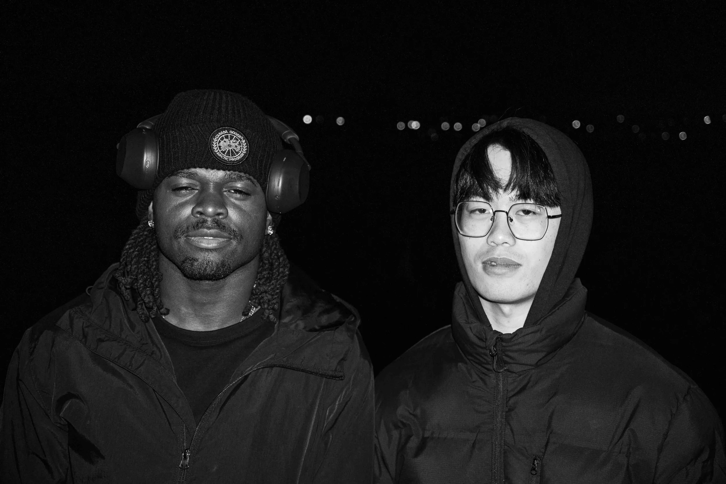 Two men standing together outdoors at night with city lights in the background. One man wears a black knit cap with headphones, a black jacket, and has dreadlocks. The other man wears glasses, a black hoodie, and a black jacket.