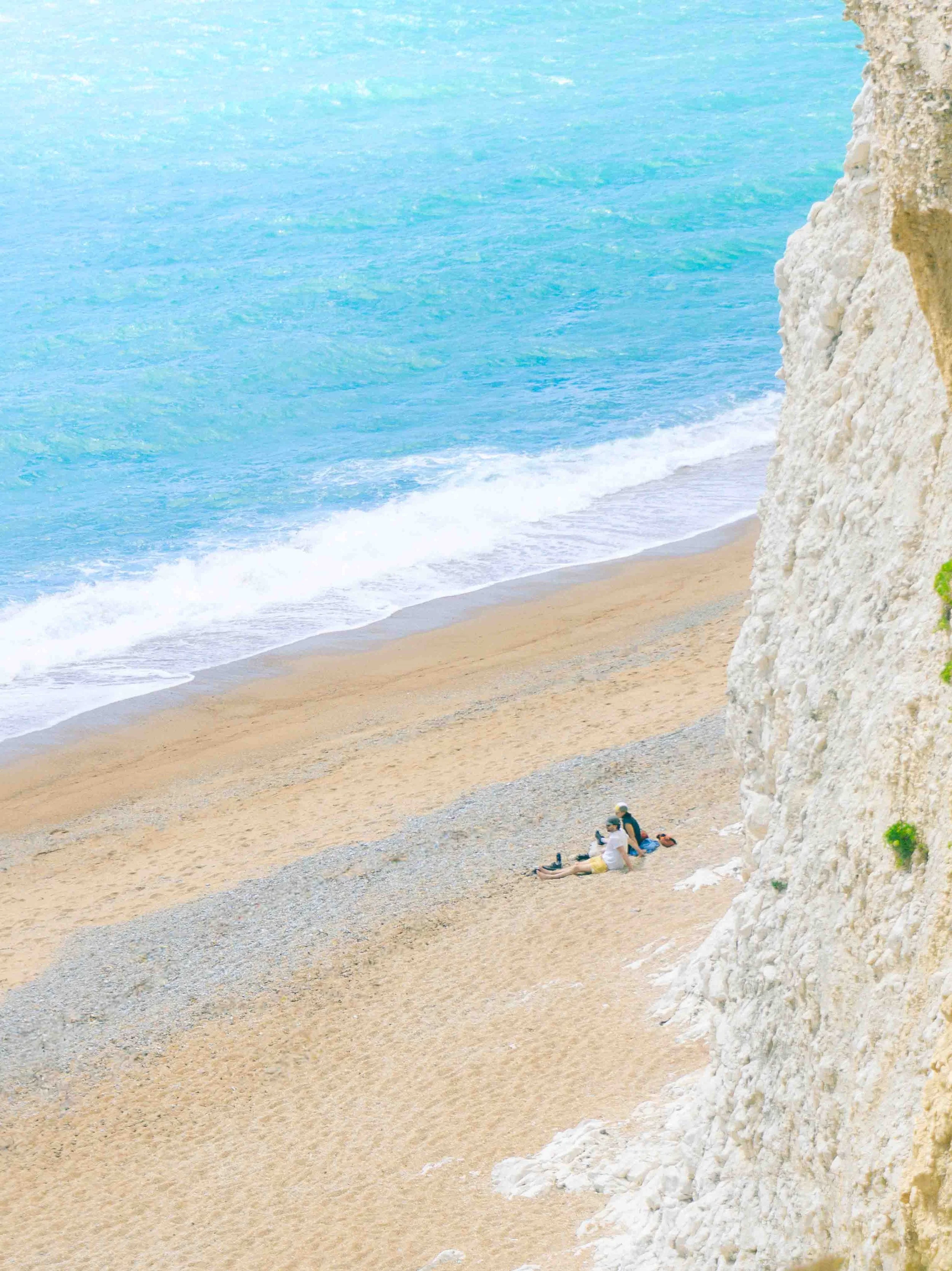 Two people sitting on sandy beach near white rocky cliff with ocean waves in the background.