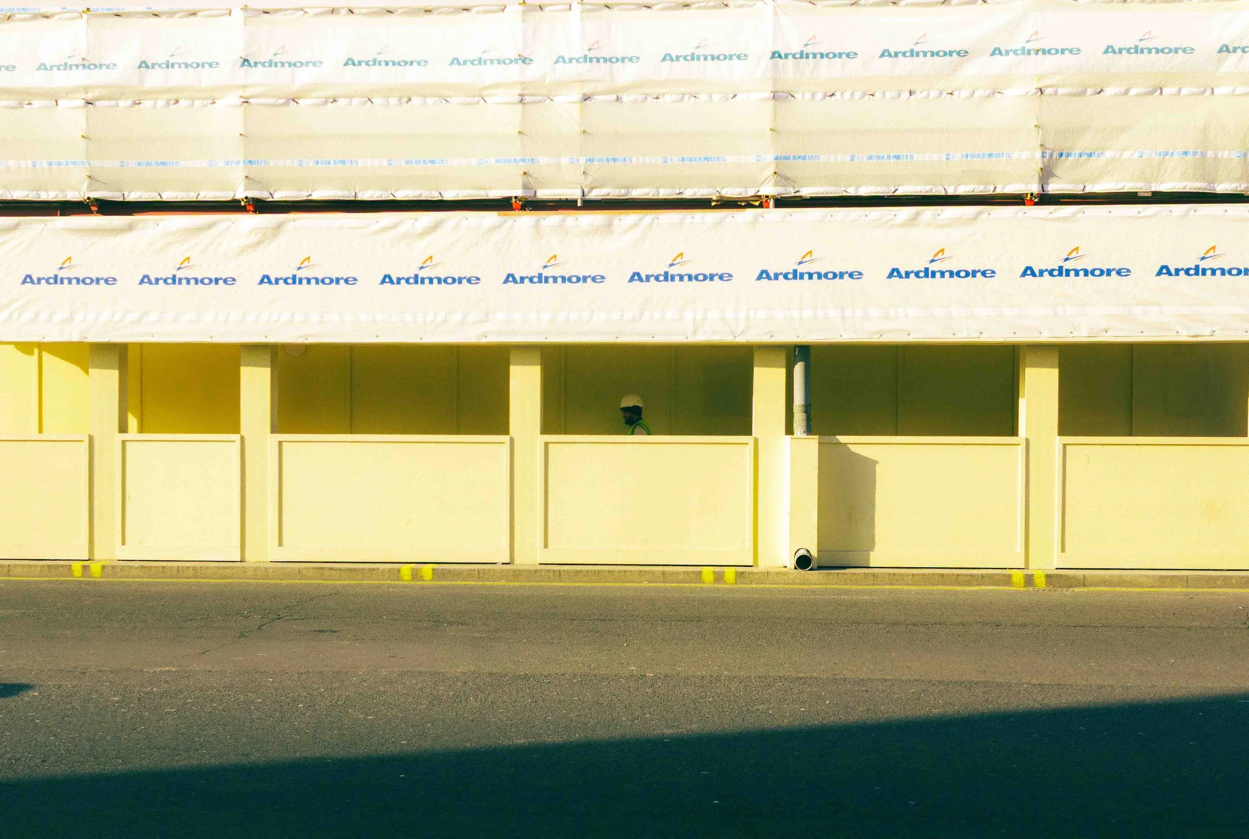 Construction site with a building covered in white tarp labeled 'Ardmore' in blue. Person wearing a hard hat and face mask visible through an opening.
