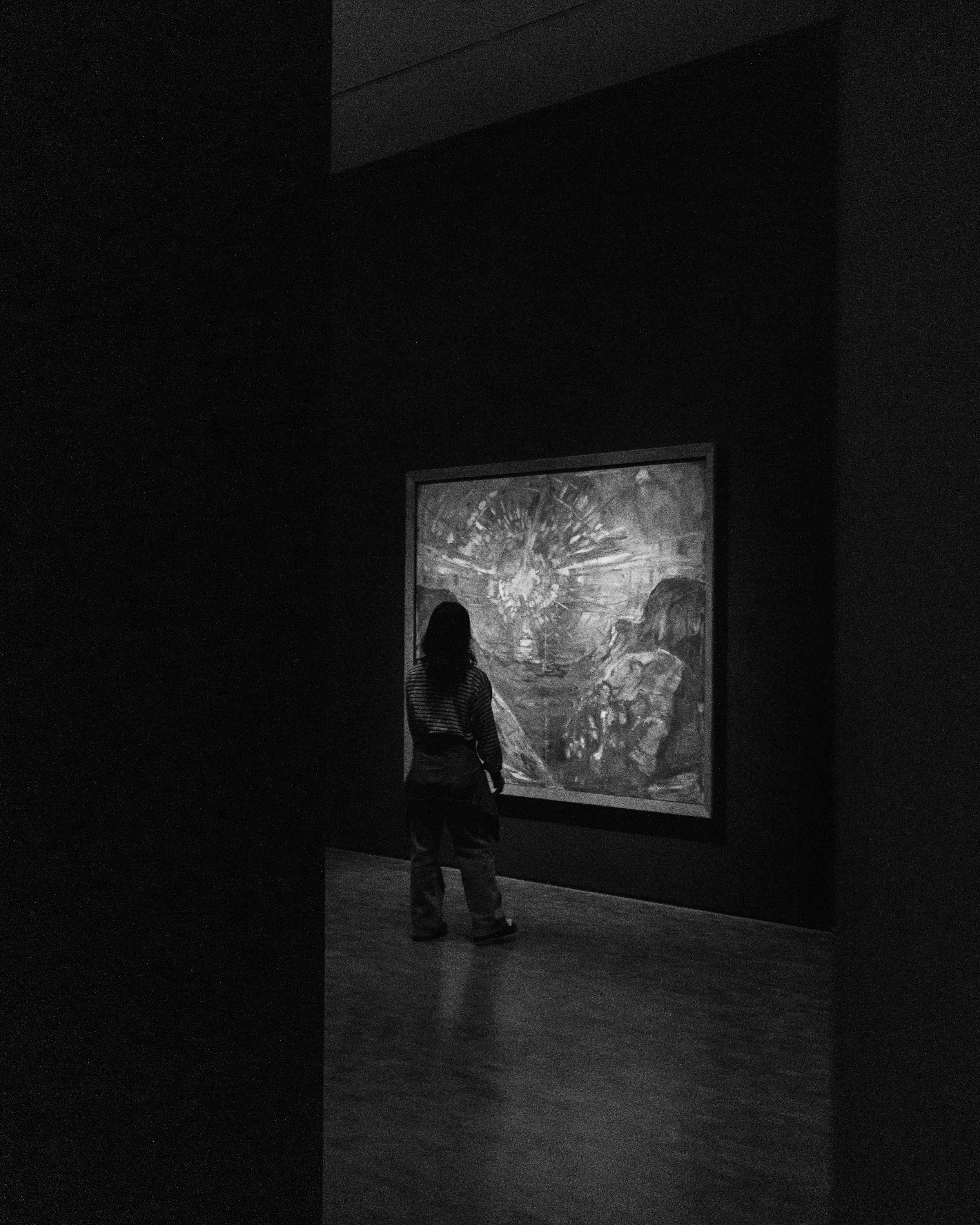 A person in a striped shirt and pants looking at a large framed painting in a dark gallery.