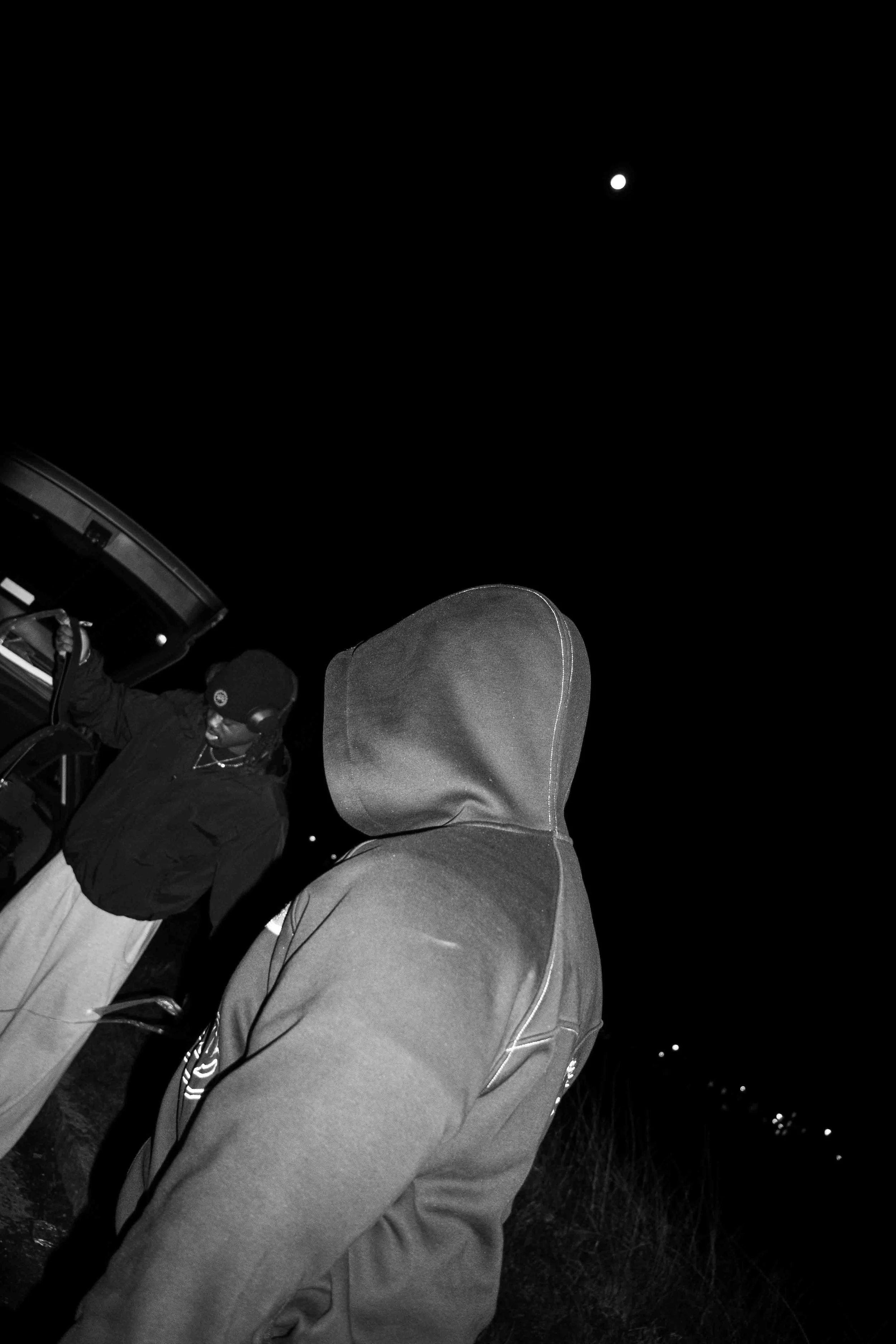 Two individuals outside at night, one with a hoodie and the other wearing a beanie and jacket, near an open vehicle trunk, with a visible moon in the dark sky.