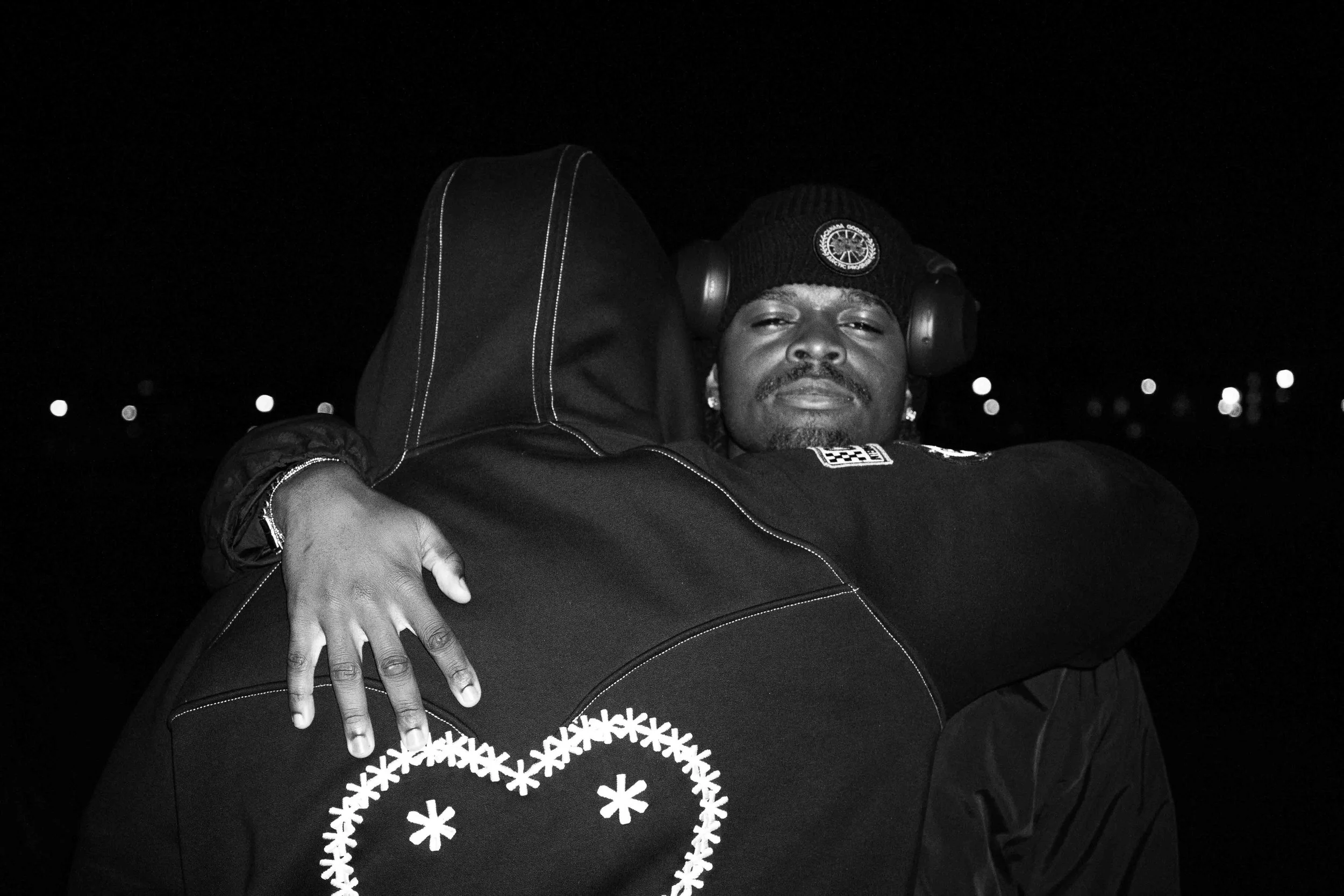 Two people hugging at night, one facing the camera with a serious expression, wearing a black beanie and headphones, the other with their back to the camera, wearing a black jacket with a graphic on the back.