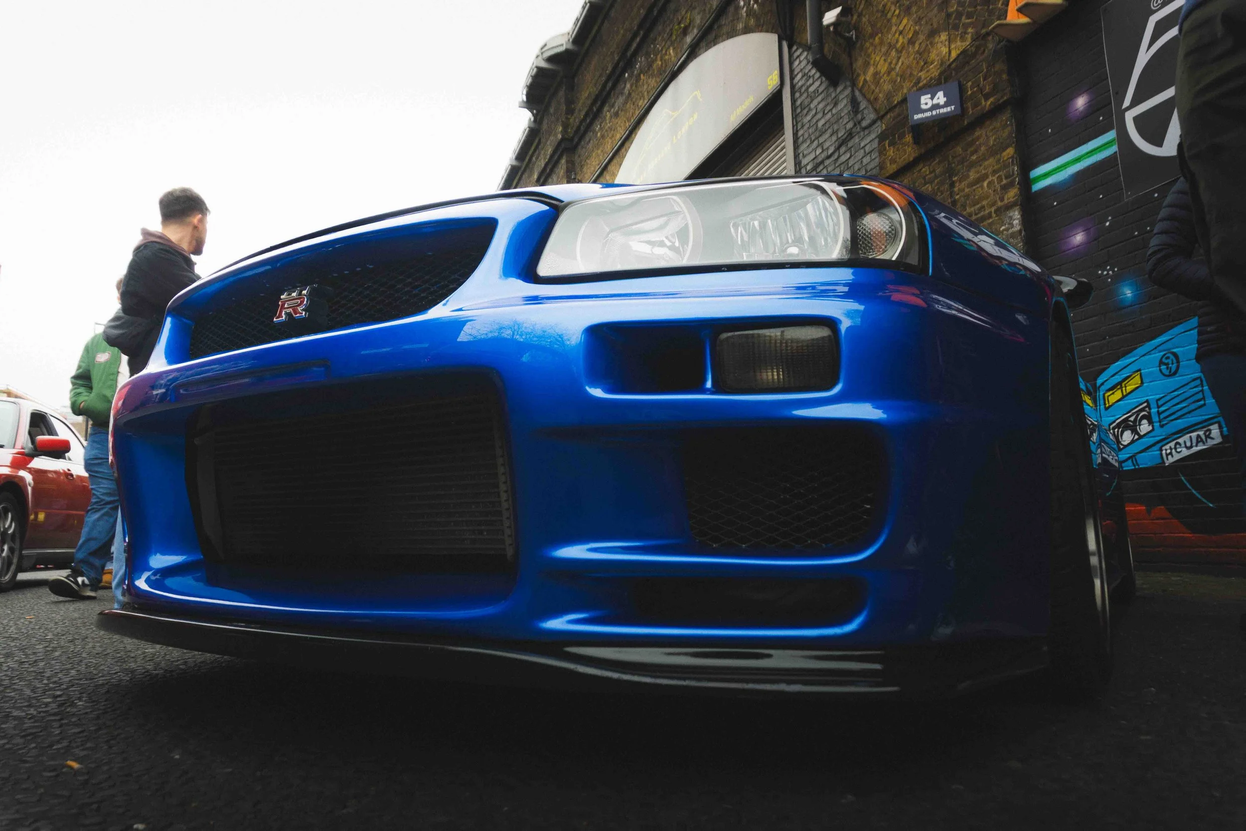 Close-up of a bright blue Nissan GT-R car parked on the street, with people standing nearby and buildings in the background.