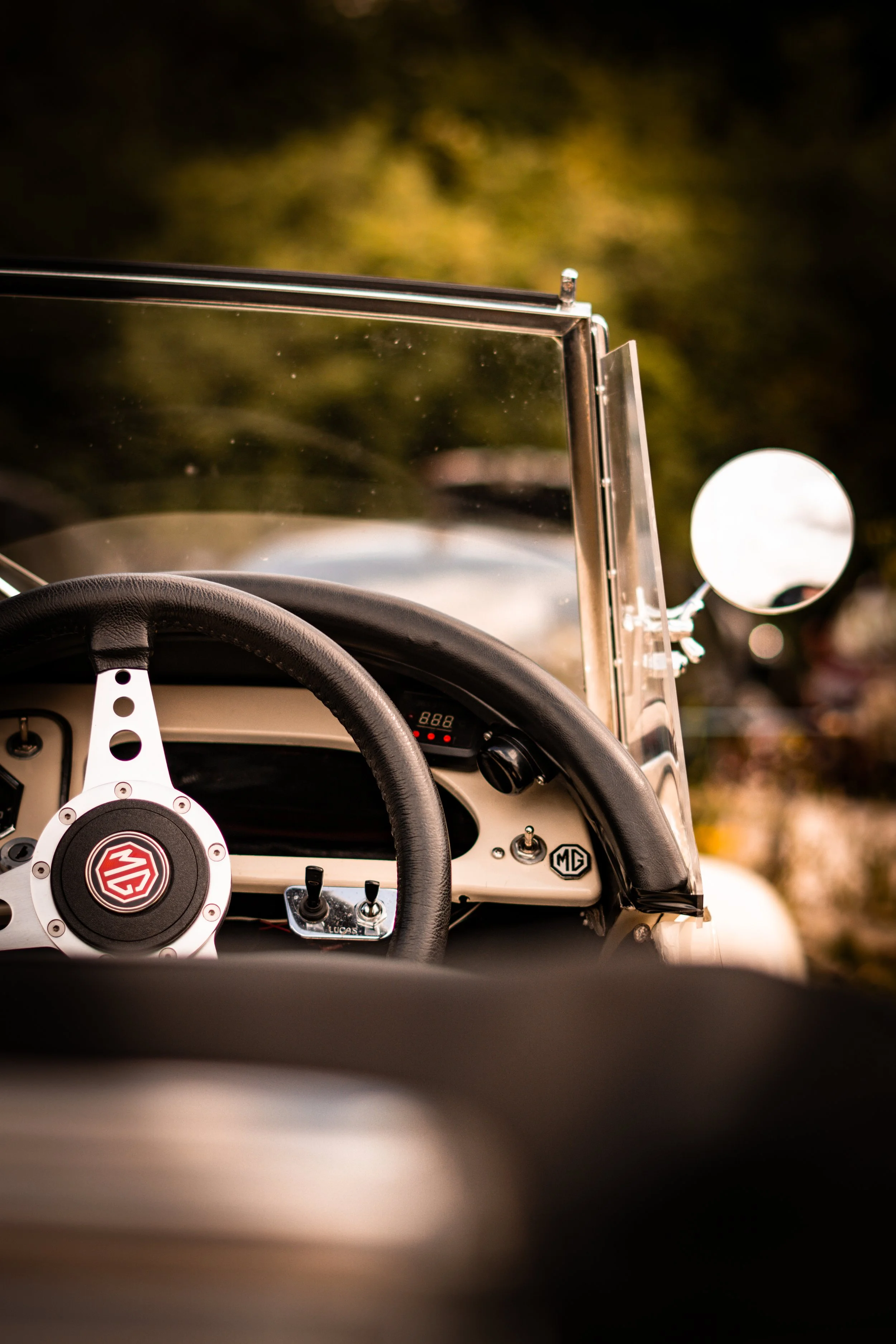 Close-up of a vintage MG car dashboard with steering wheel, gauges, and side mirror visible, outdoors during daytime.