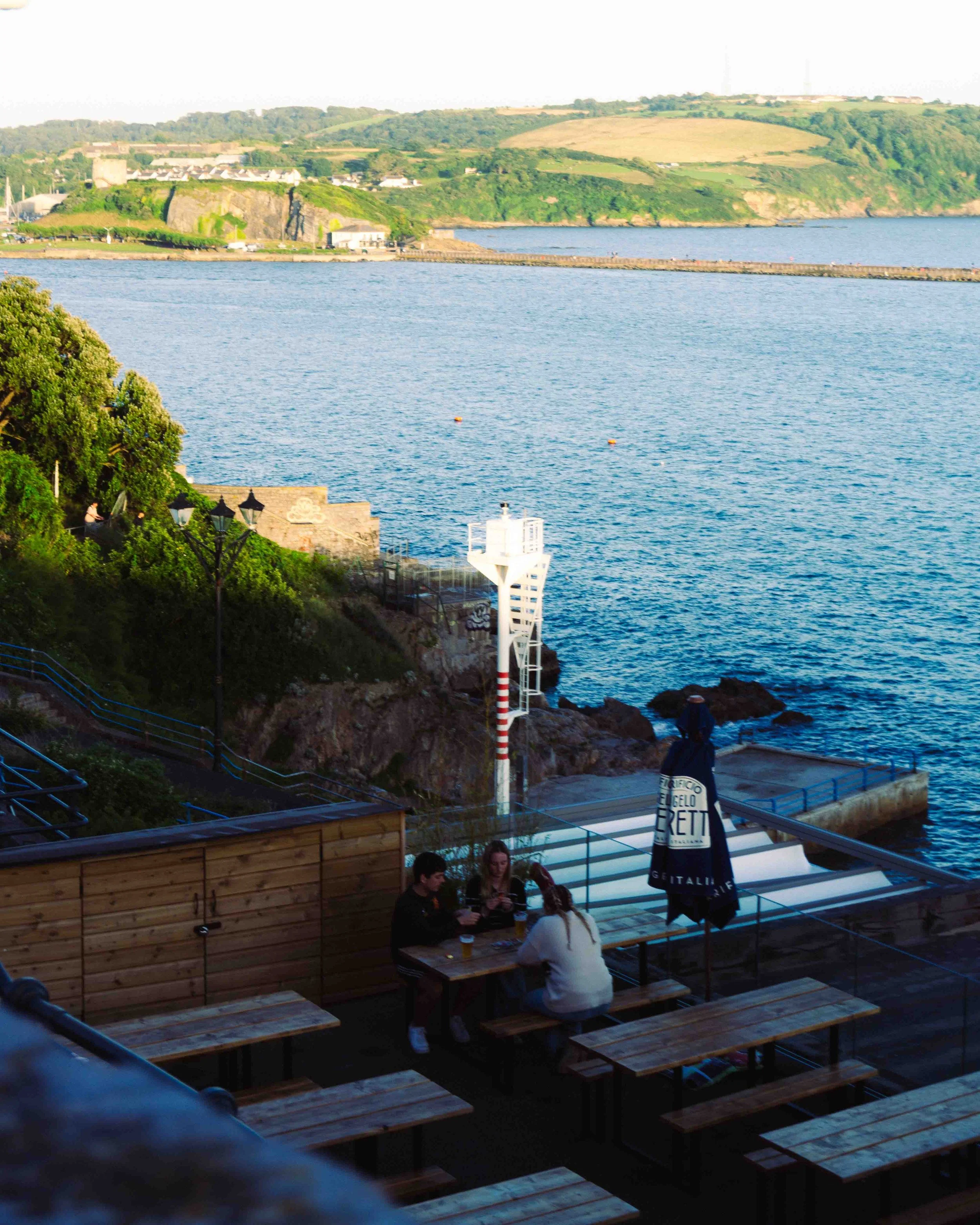 People sitting at wooden tables on an outdoor deck overlooking the water, with a lighthouse and green hills in the background.